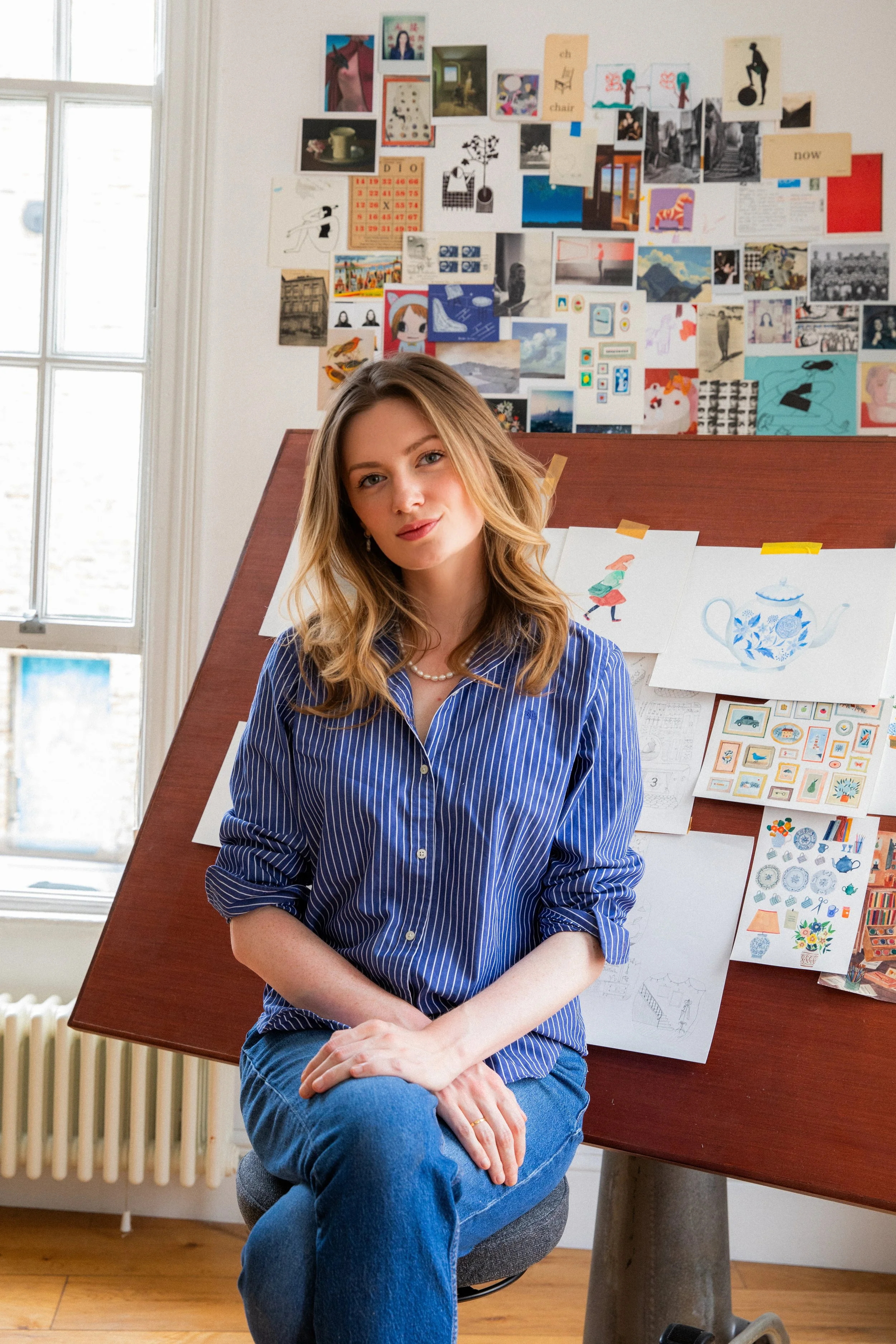 A young woman with wavy blonde hair wearing a blue striped shirt and jeans, sitting on a gray chair in a room with natural light. Behind her is a bulletin board filled with various colorful pictures, drawings, and notes, with a window to the left and a wooden floor.