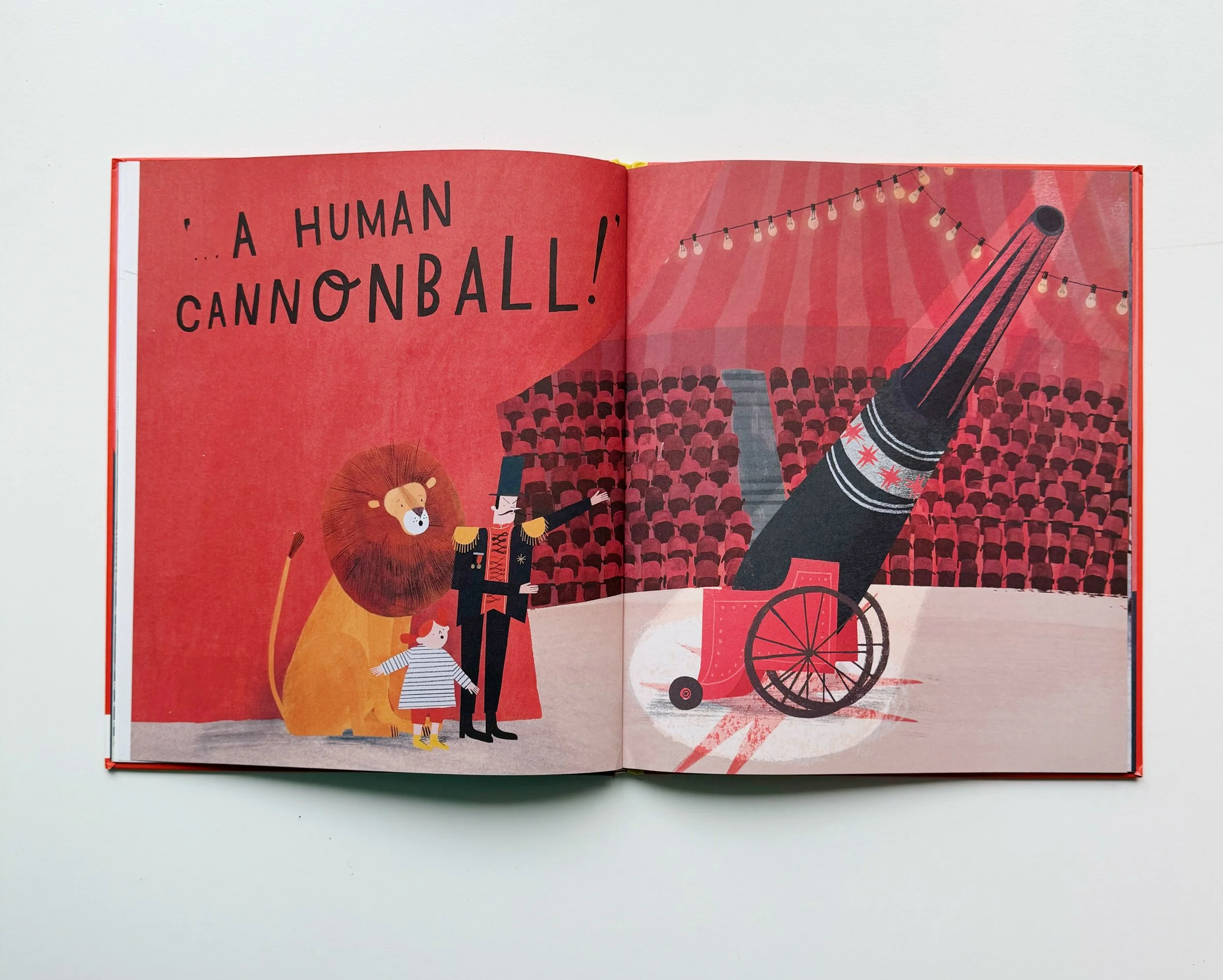 Open illustrated children's book showing a circus scene, with text reading 'A HUMAN CANNONBALL!' and depicting a cannon, a lion, a circus ringmaster, and children watching.