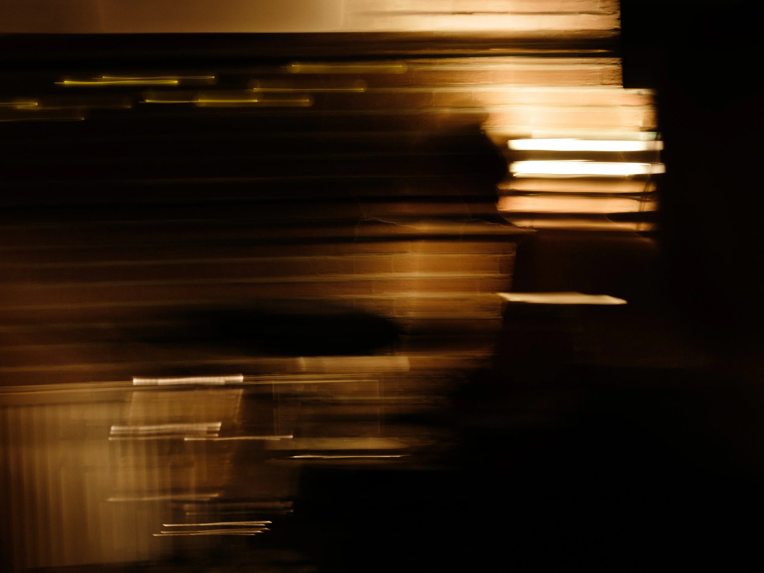 A blurry, dark image with streaks of light, featuring a silhouette of a person standing near a window with blinds, illuminated by a warm glow.