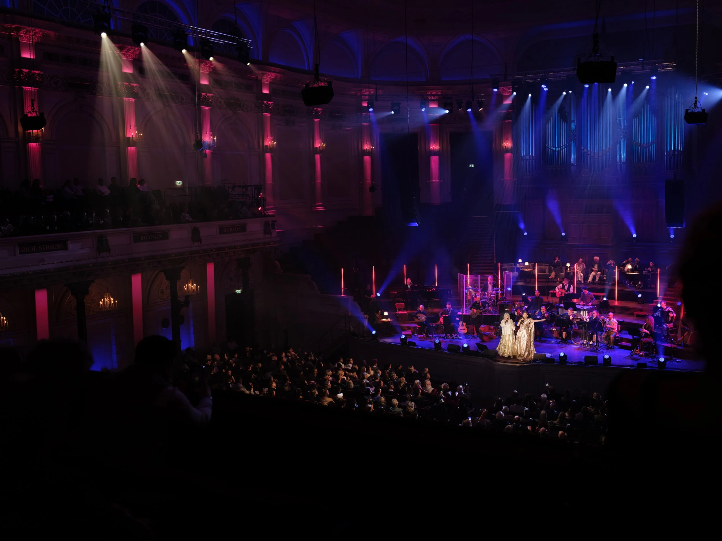 A stage with a musical performance, including two singers in golden dresses, surrounded by musicians, with colorful spotlights and pink lighting in a large concert hall filled with an audience.