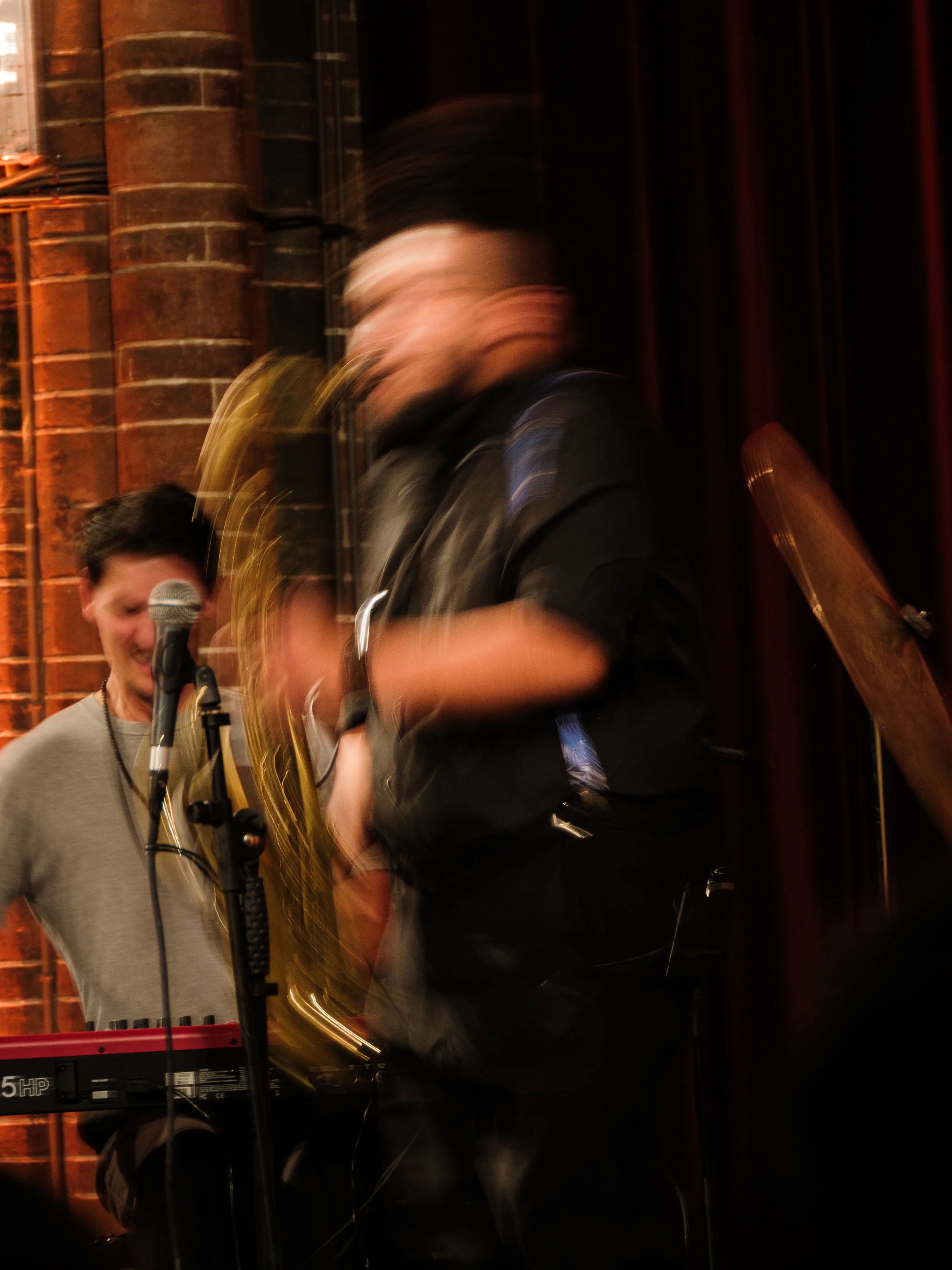 Blurred image of two musicians performing on stage, one playing a saxophone and the other at a keyboard, with a brick wall background and dark curtain.