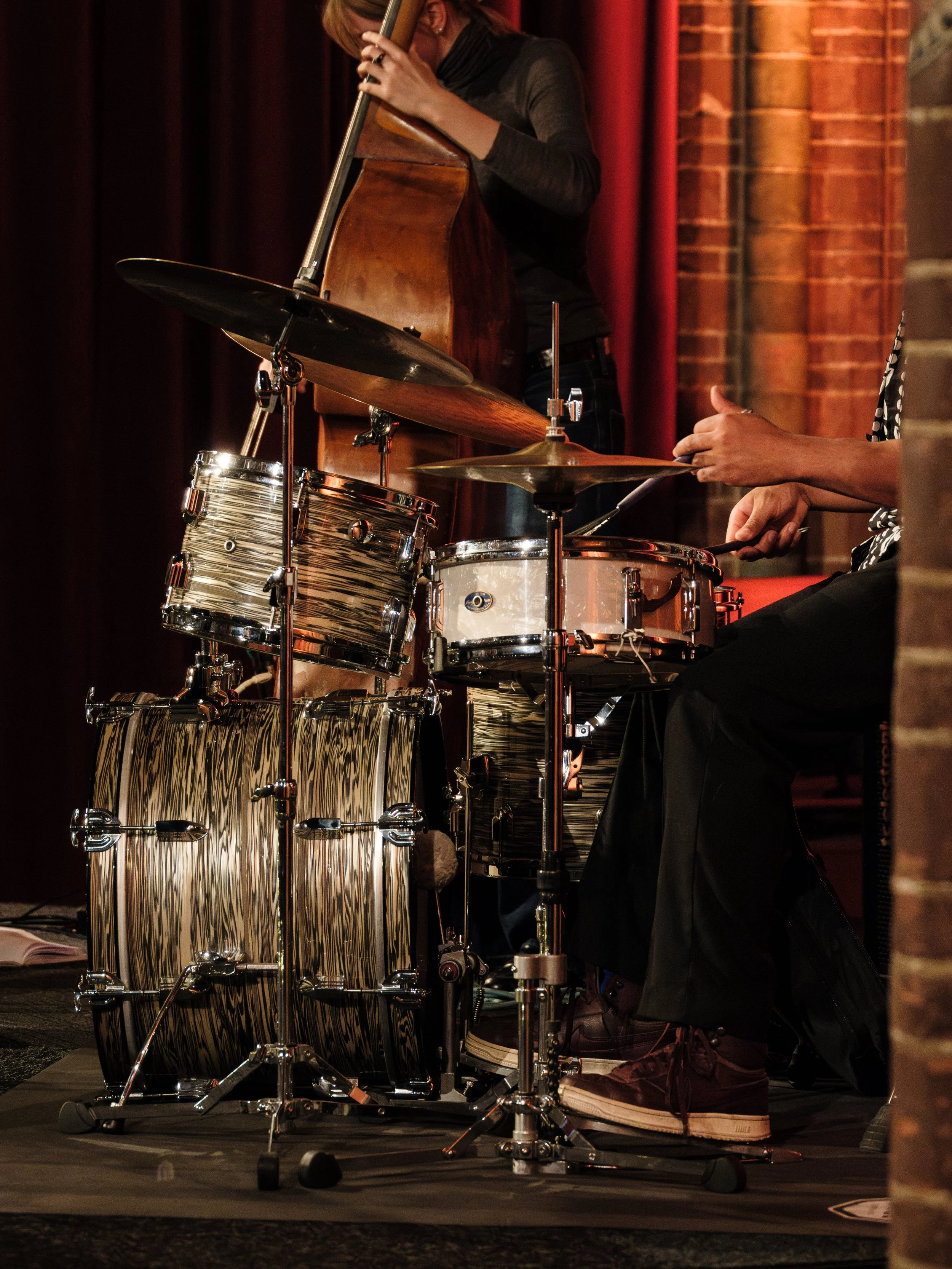 Close-up of musicians playing percussion instruments and a double bass on stage, with brick wall background and dark curtains.