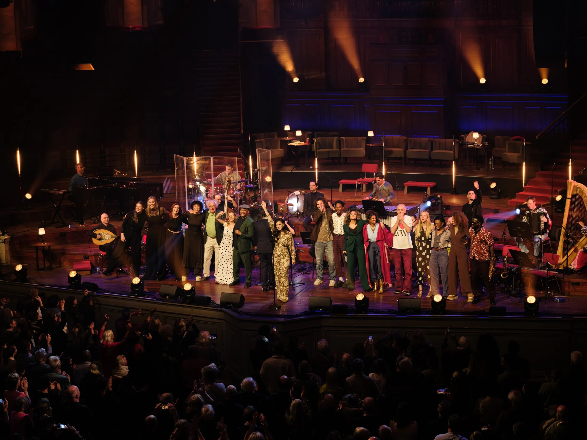 Musicians and performers on stage after a concert, with an audience in front, in a theater with wooden paneling and warm lighting.