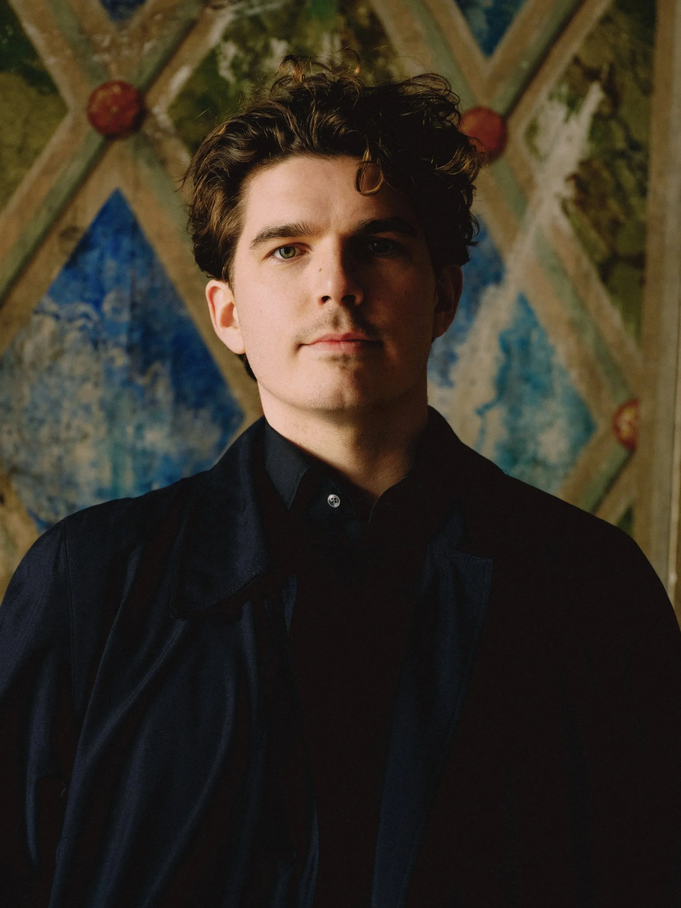A young man with wavy brown hair and light stubble, dressed in a dark jacket and shirt, stands in front of a painted backdrop featuring geometric patterns and landscape elements.