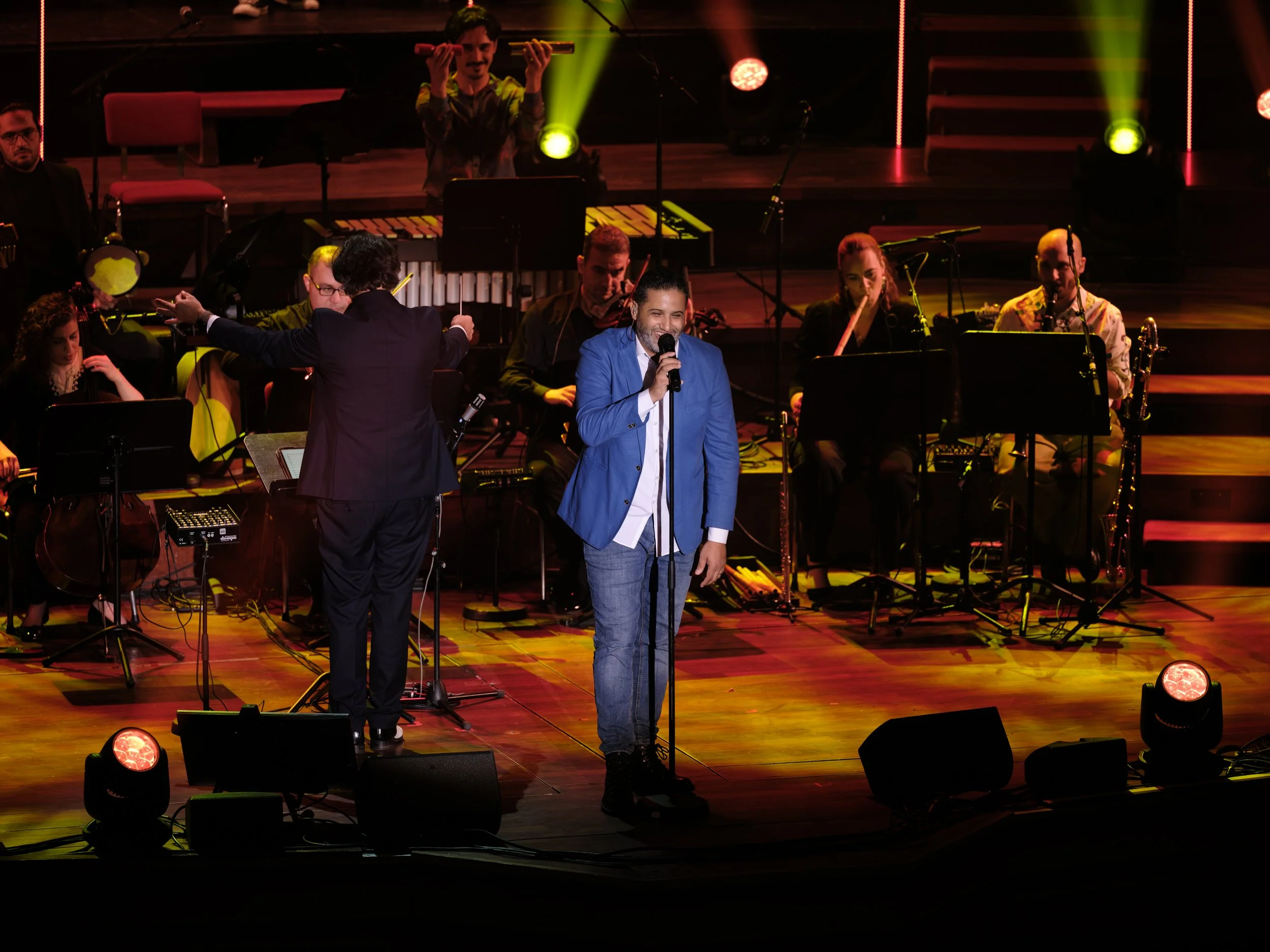 Male singer in a blue blazer performing with a band on stage with colorful lighting.