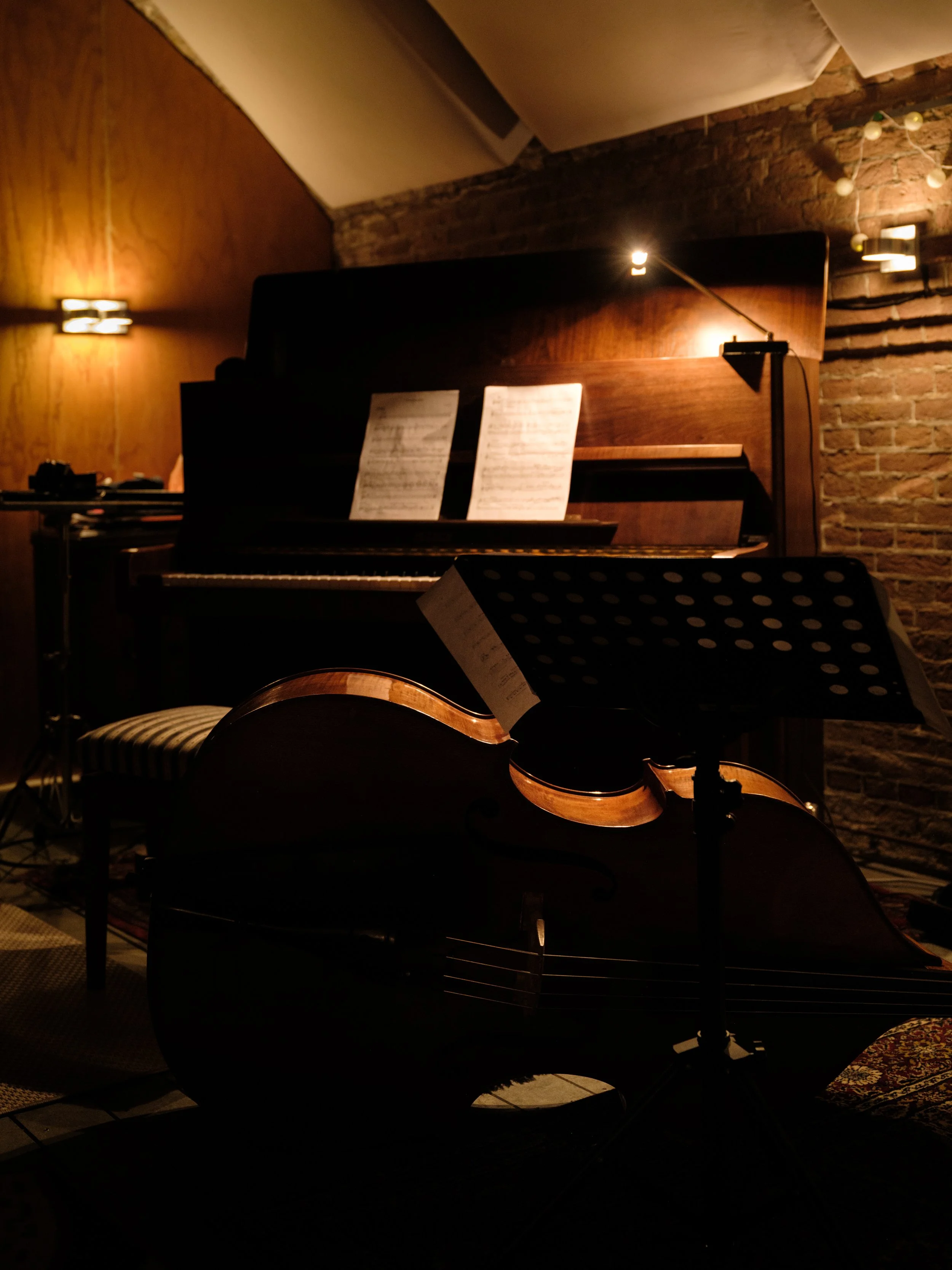 A dimly lit room with a piano, sheet music, a music stand, and a cello with a sheet on it, set against a brick wall and wooden wall paneling.