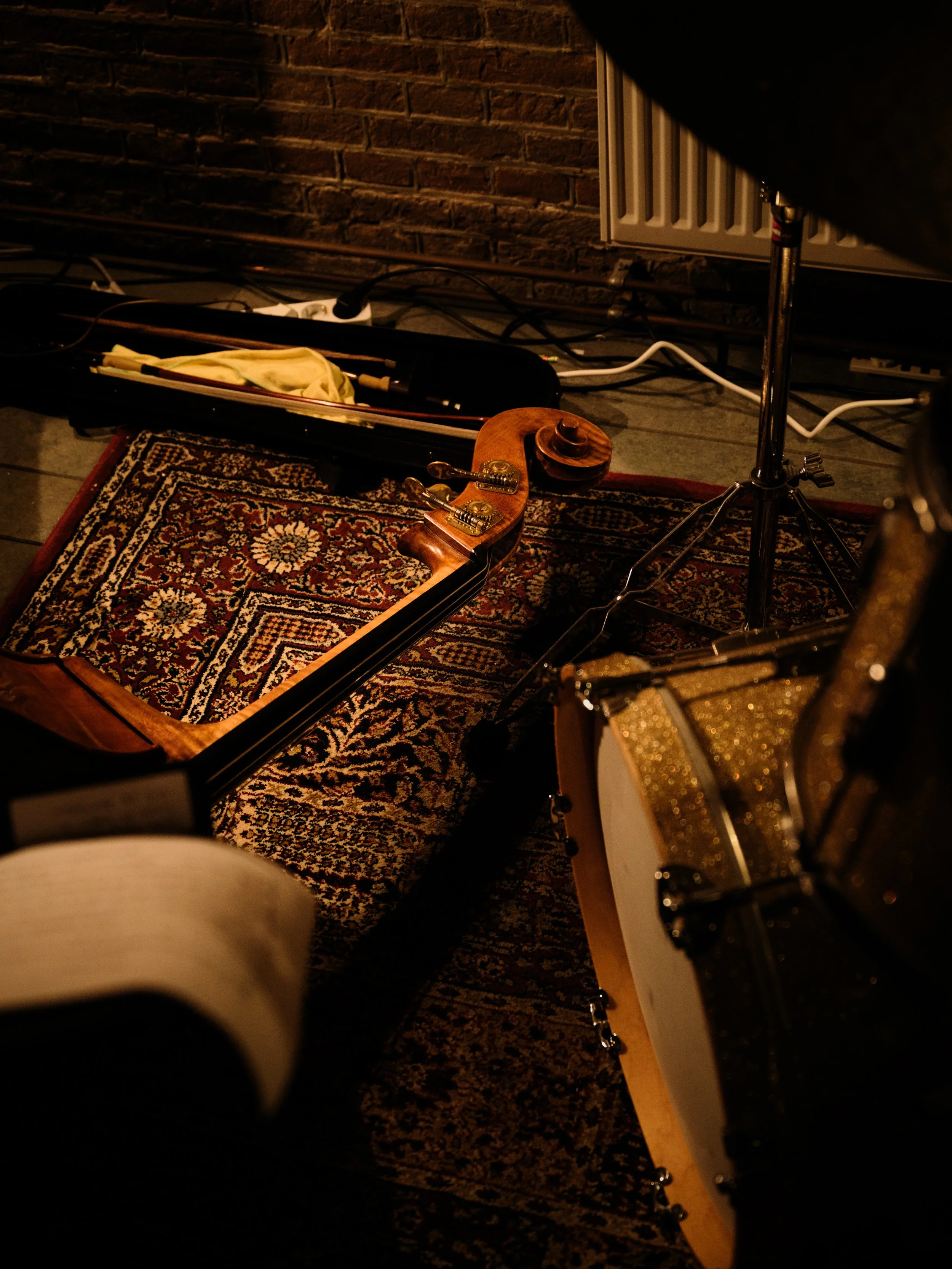 Musical instruments including a violin lying on a patterned rug, a drum kit, and a music stand in a dimly lit room with cables and a brick wall in the background.