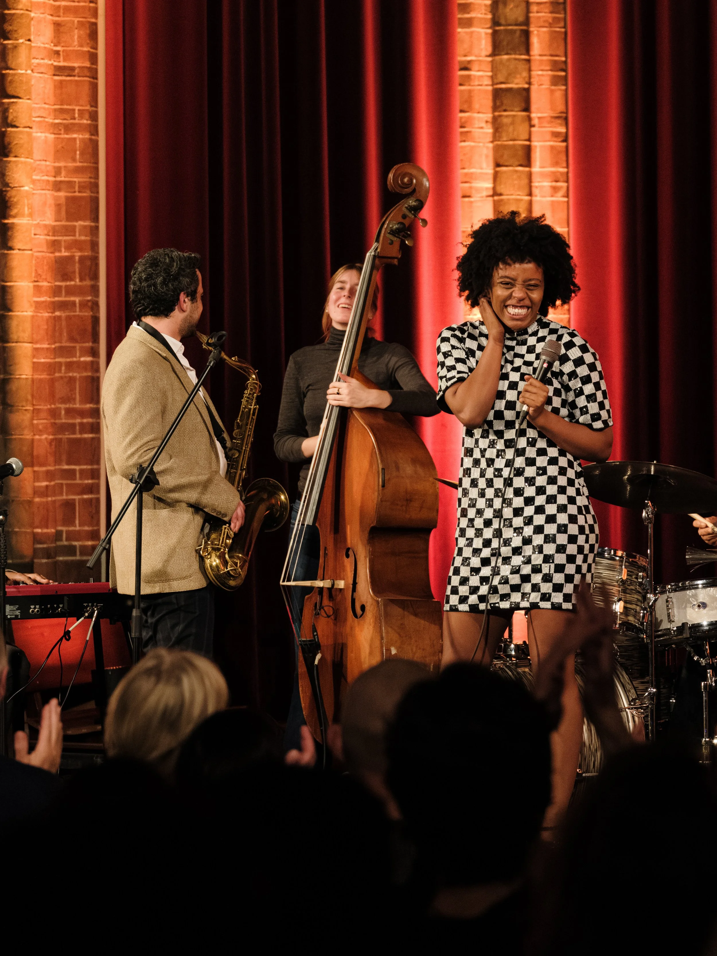 Jazz band performing on stage with a woman singing into a microphone, backed by musicians playing a double bass, saxophone, keyboard, and drums, in a venue with red curtains and brick walls.