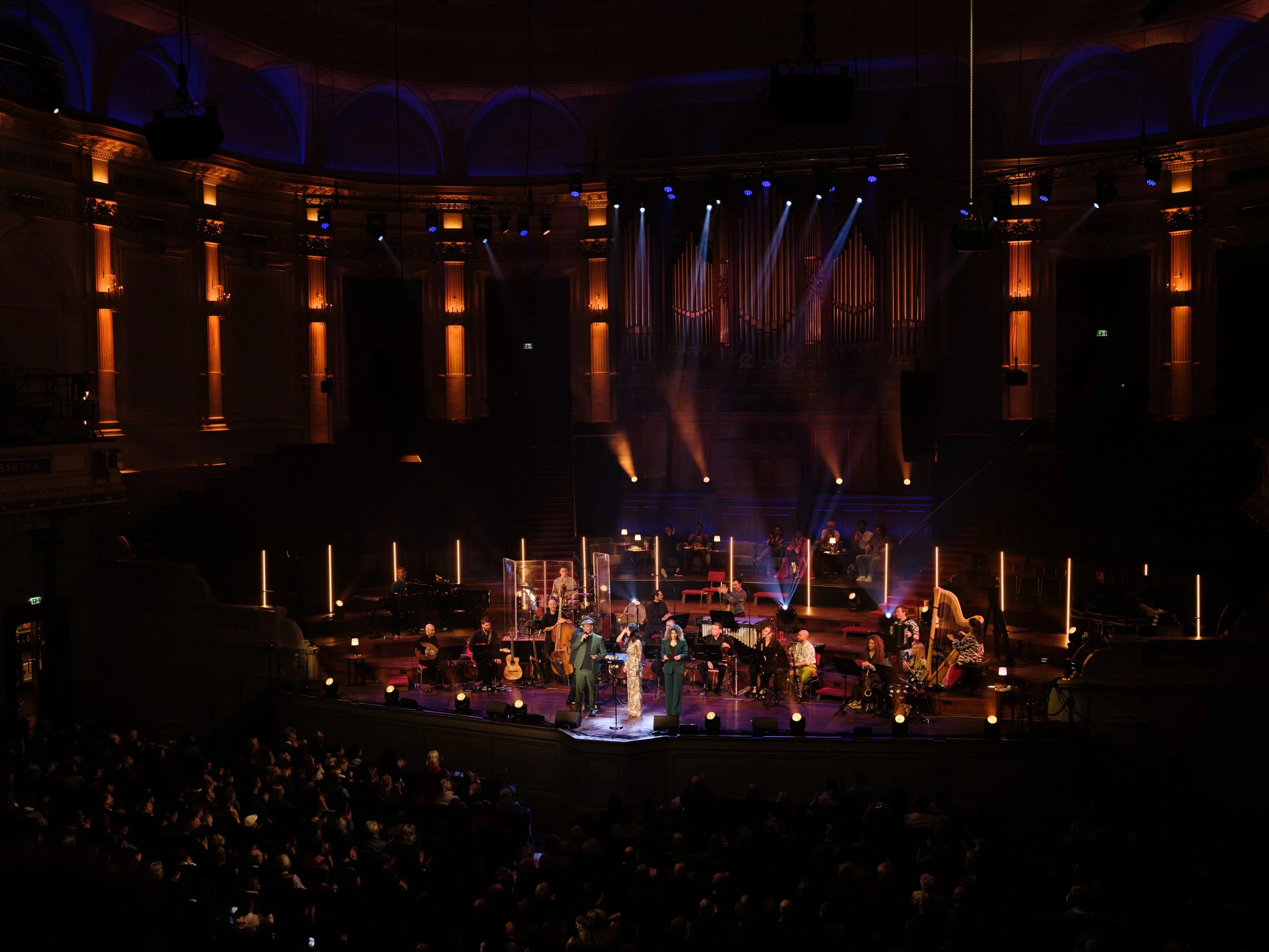 Live concert on a stage in a grand concert hall with musicians and singers, illuminated by stage lights, with an audience watching.