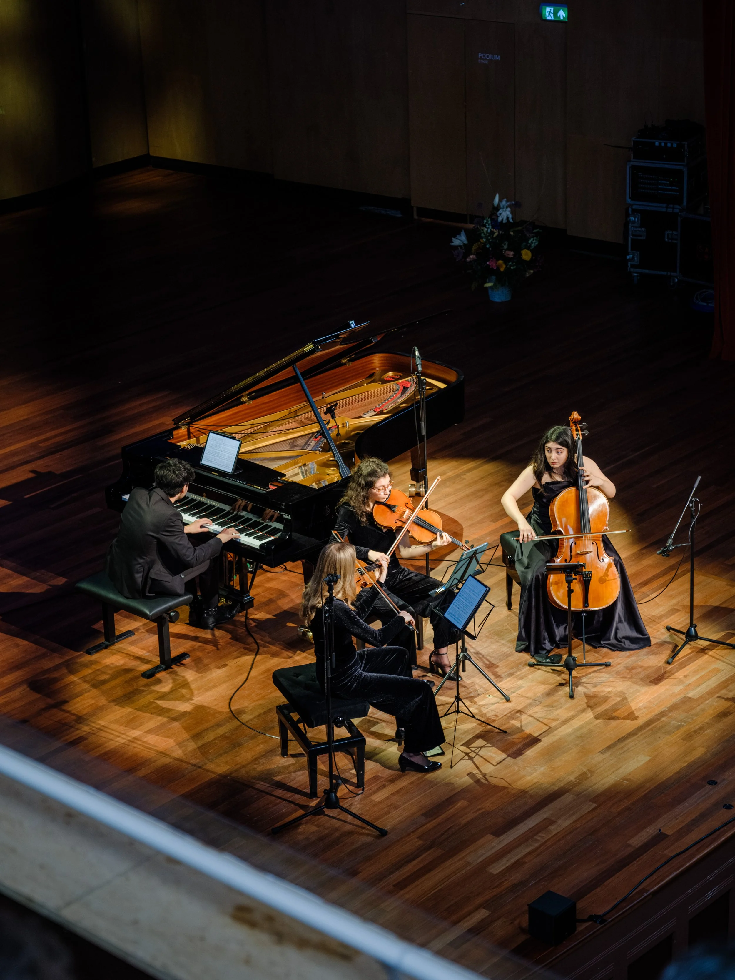 A classical music ensemble performing on stage with a grand piano, violin, and cello, in a concert hall.