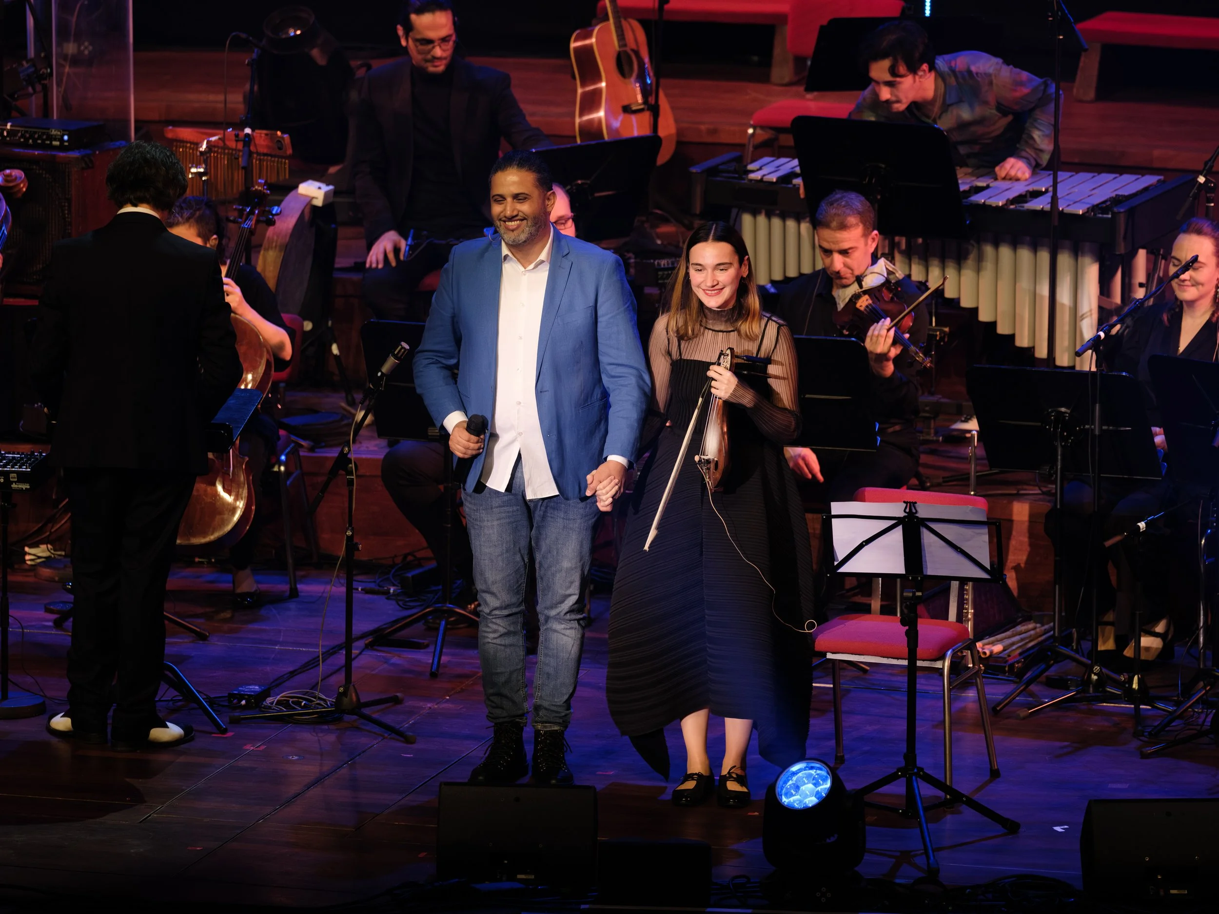 Musicians and performers on stage after a concert, including a man in a blue jacket holding a microphone, a woman with a violin, and orchestra members with various instruments.