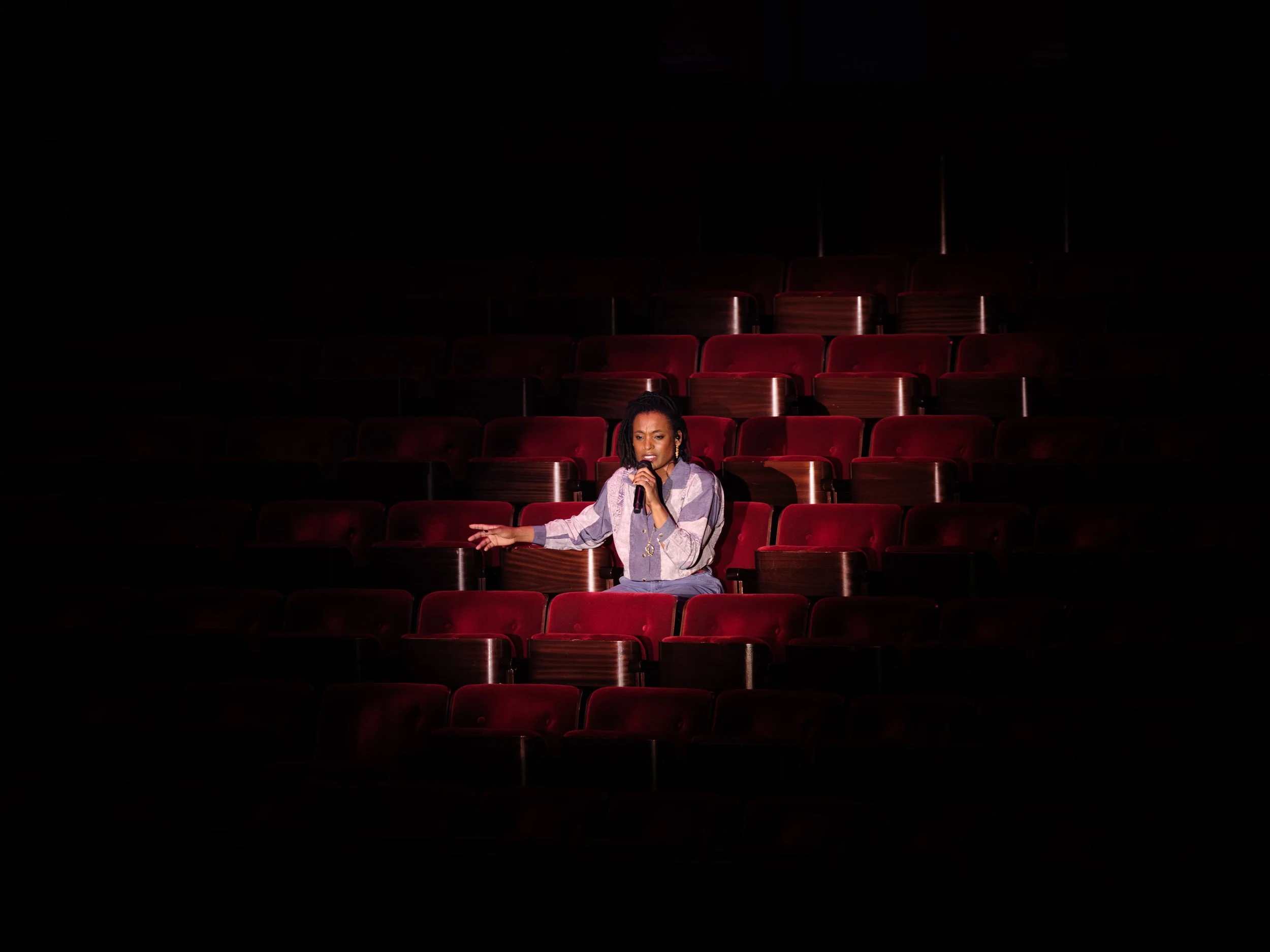 A woman singing into a microphone in an empty theater with red seats.