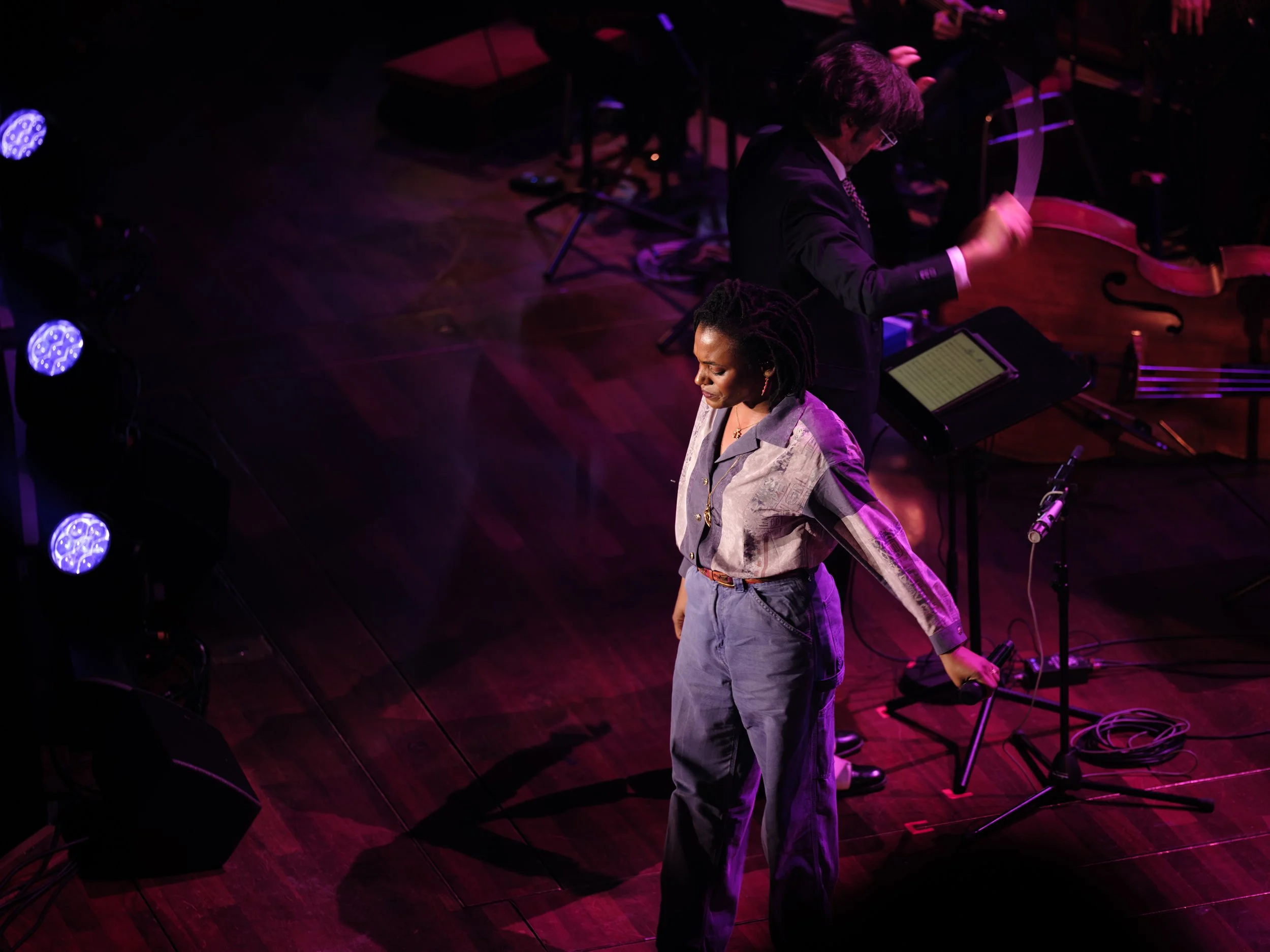 A woman stands holding a microphone on a stage with musical instruments and a man playing a violin in the background, under stage lighting.