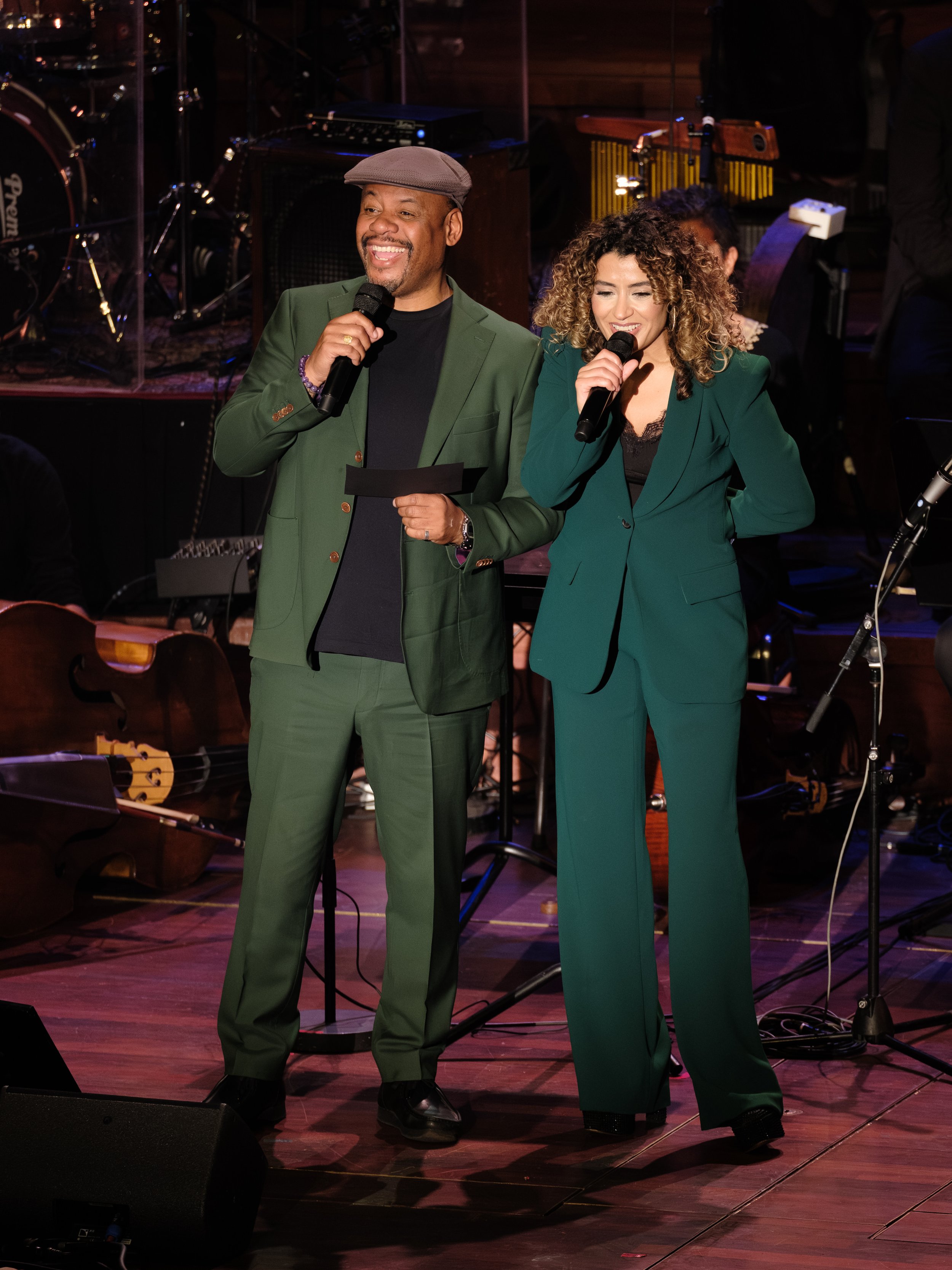 Two people in green suits standing on stage, holding microphones, smiling, and speaking to an audience during a live event with musical instruments in the background.