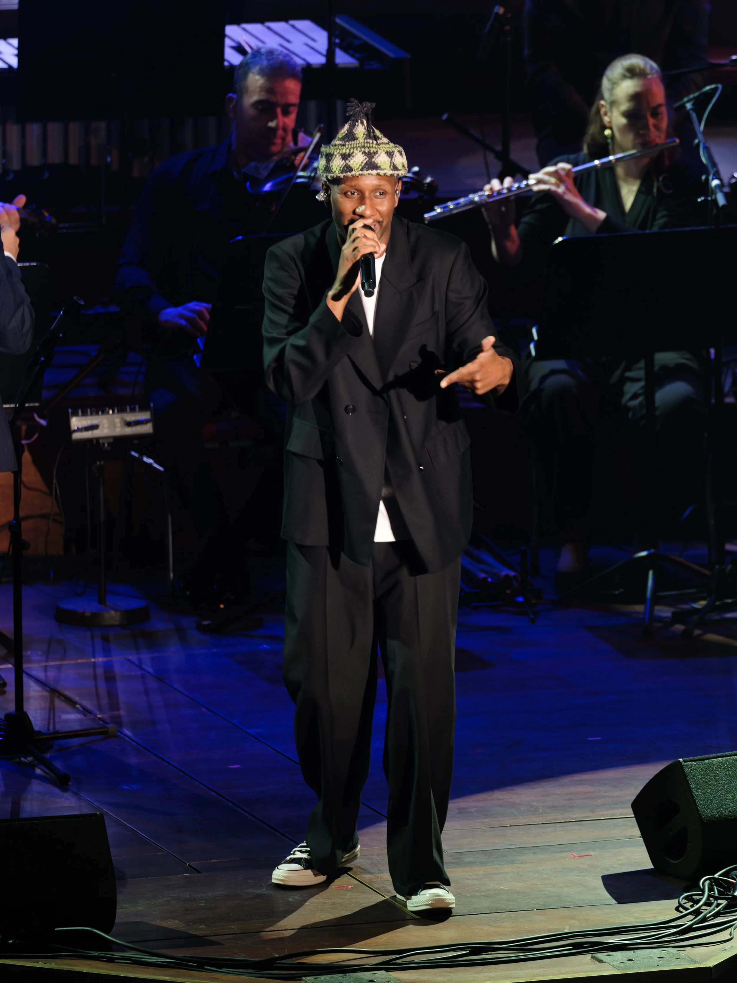 A performer in black clothing and a patterned beanie is singing into a microphone on stage, with an orchestra in the background.