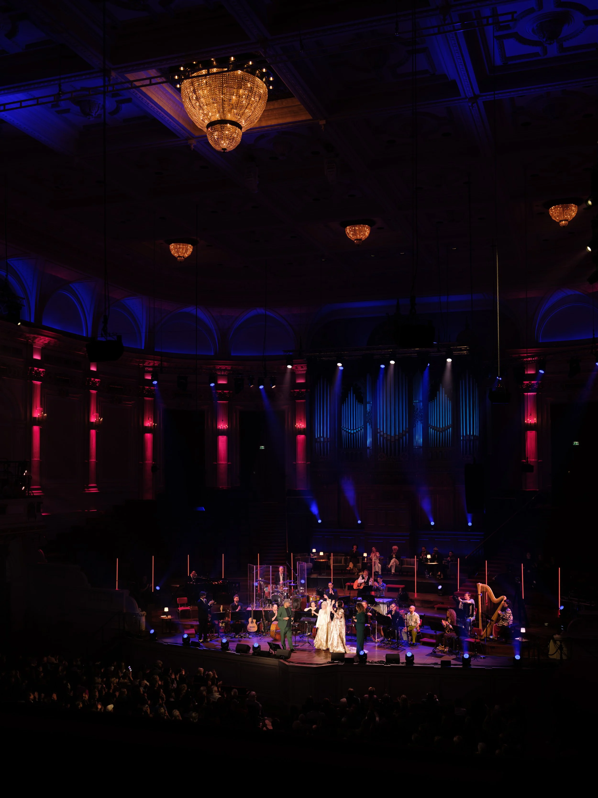 Musicians perform on stage in a theater with ornate decor, chandeliers overhead, colorful lighting, and a seated audience.