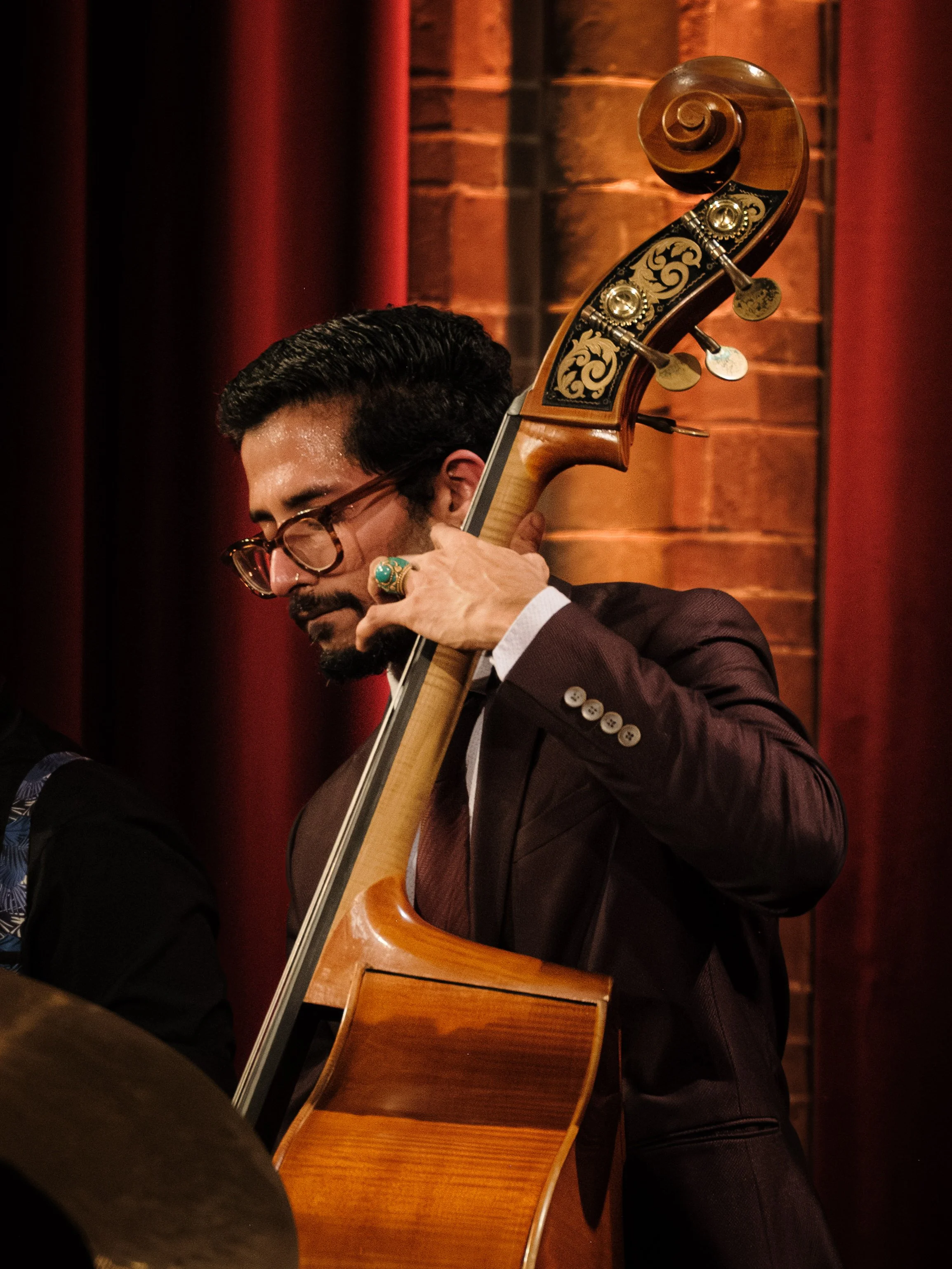 A man with glasses and a mustache playing a double bass in a dimly lit room with brick walls and red curtains.