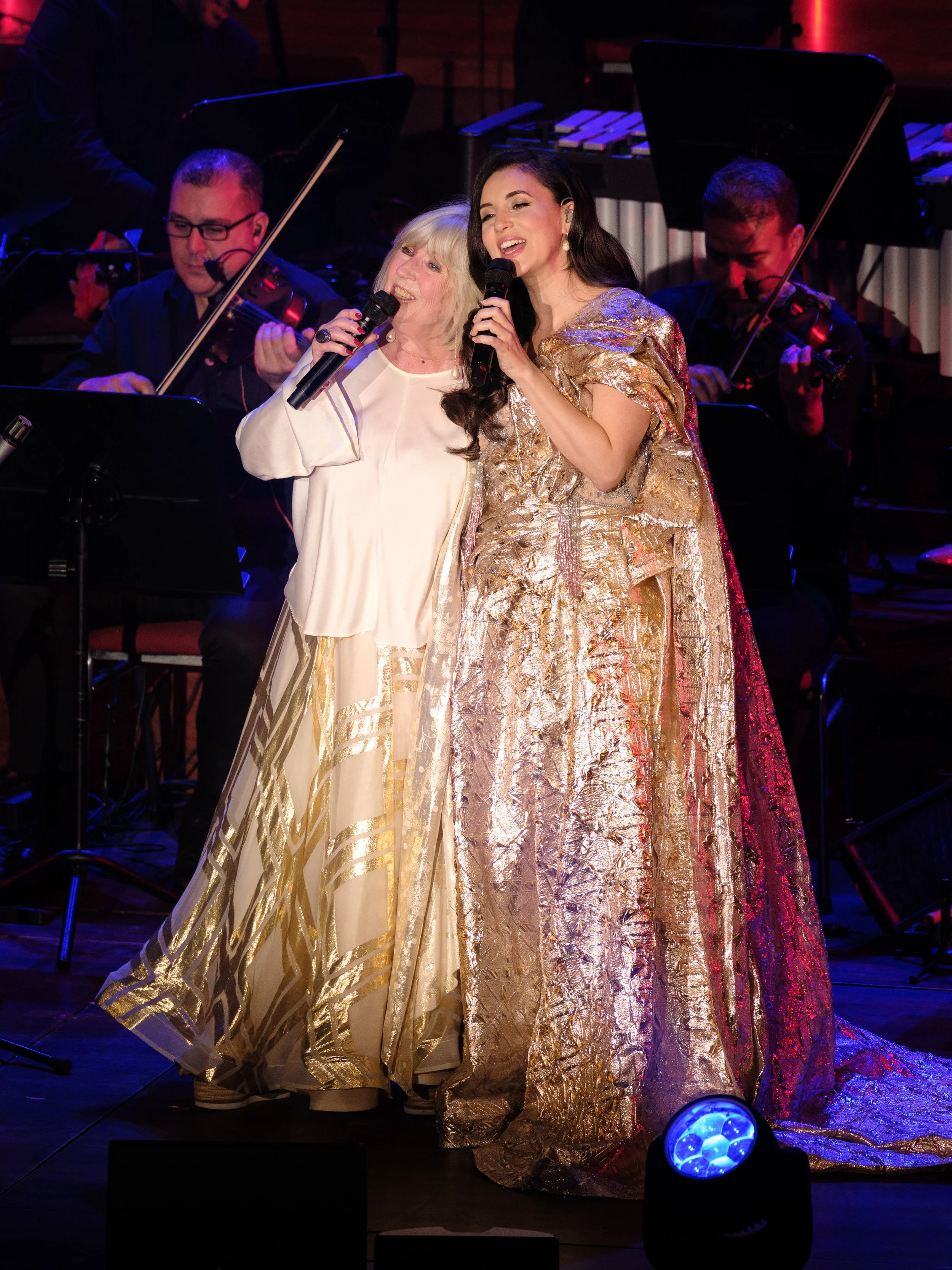 Two women singing on stage with a live orchestra behind them, wearing elegant gold and cream dresses.