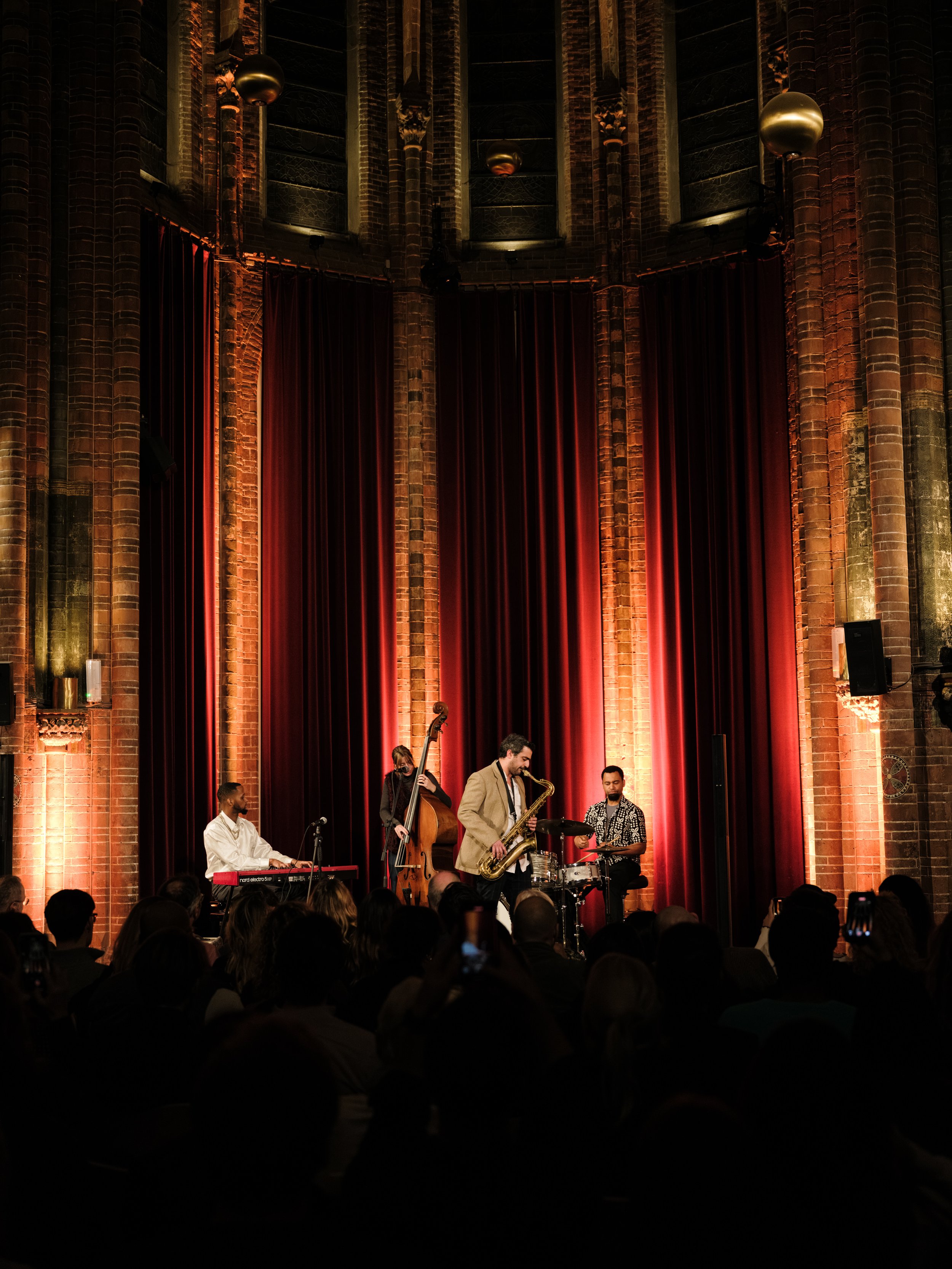 Jazz band performing on stage in front of an audience inside a brick-walled venue with tall, arched windows and red curtains.