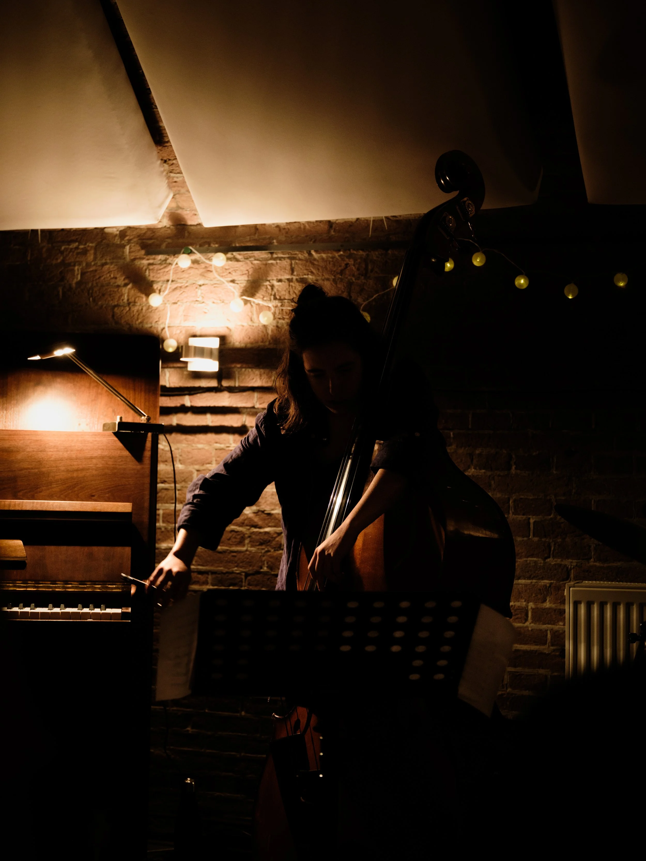 A woman adjusting sheet music on a stand in a dimly lit room with a brick wall, a piano, and string lights in the background.