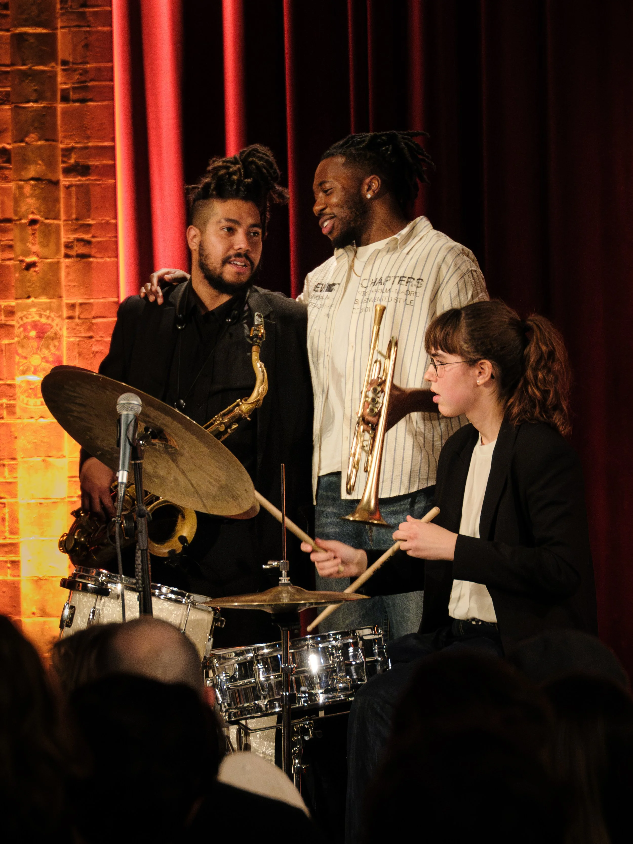 Three musicians performing on stage: a drummer, a saxophonist, and a trumpeter, with an audience watching.