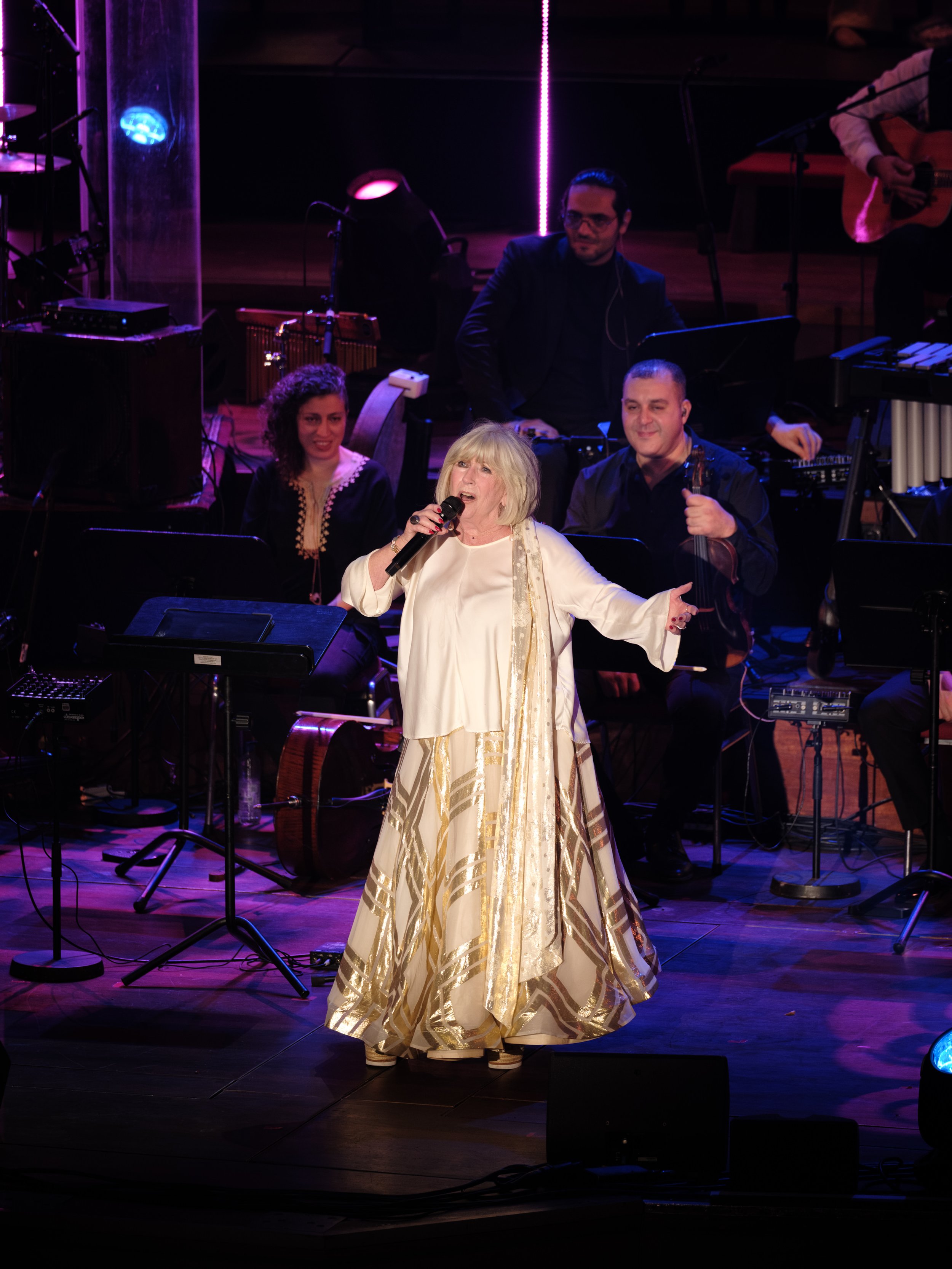An elderly woman in a white and gold dress sings into a microphone on stage, accompanied by a live band playing musical instruments. The scene is illuminated with stage lights.