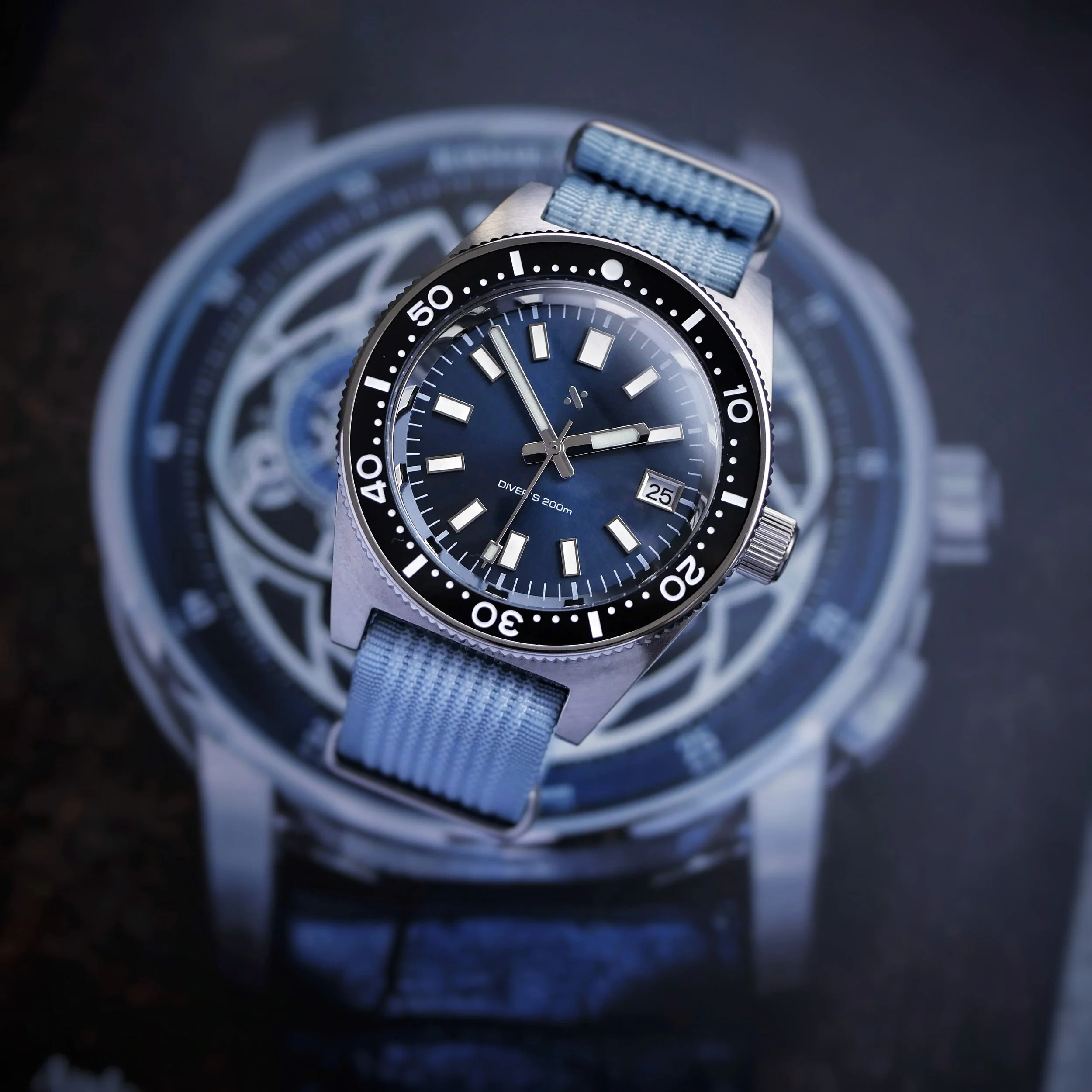 Blue-dial Namoki NMK07 with a pale blue woven strap resting over a printed watch image.