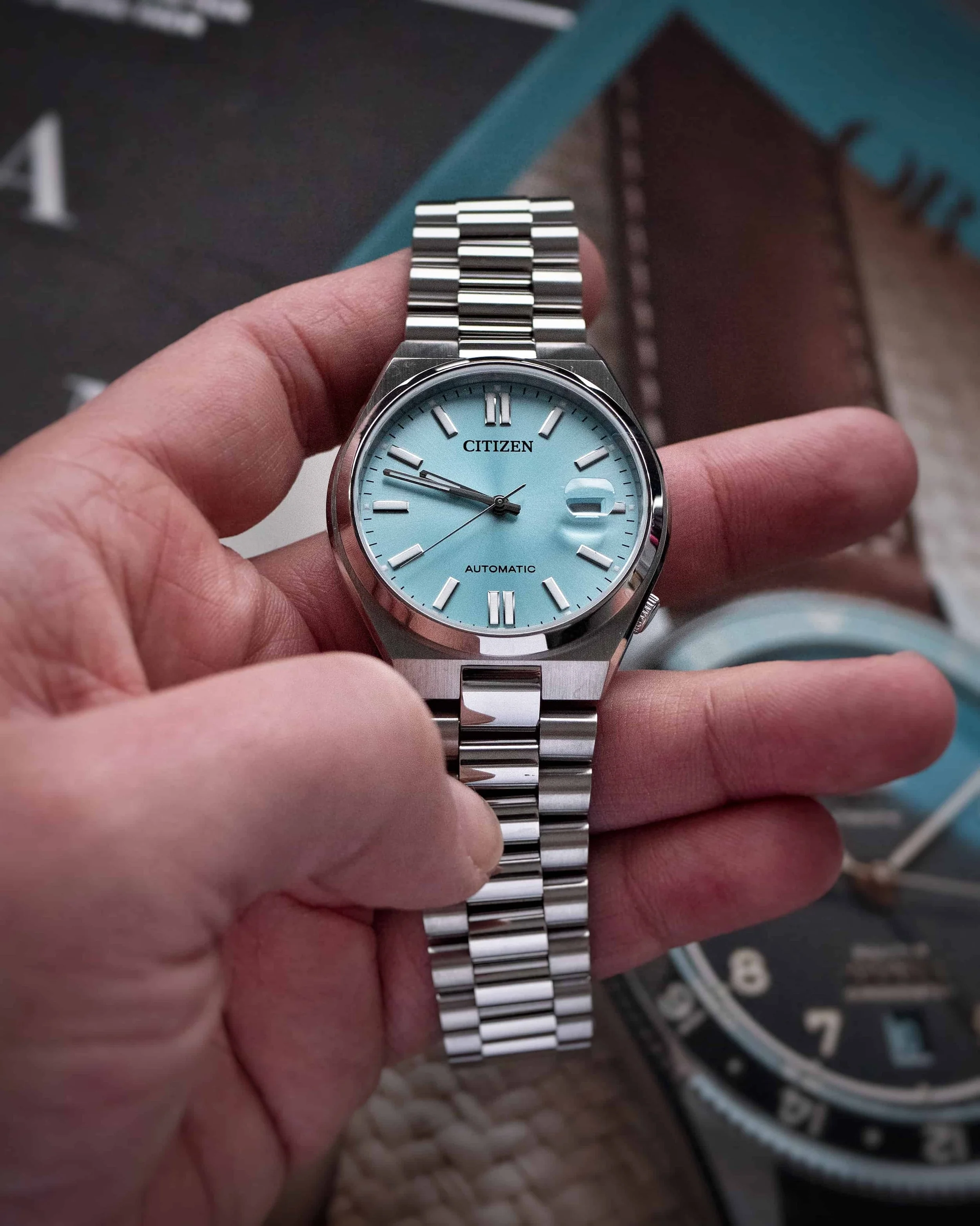 Turquoise-blue Citizen Tsuyosa automatic held in hand with its stainless-steel bracelet extended.