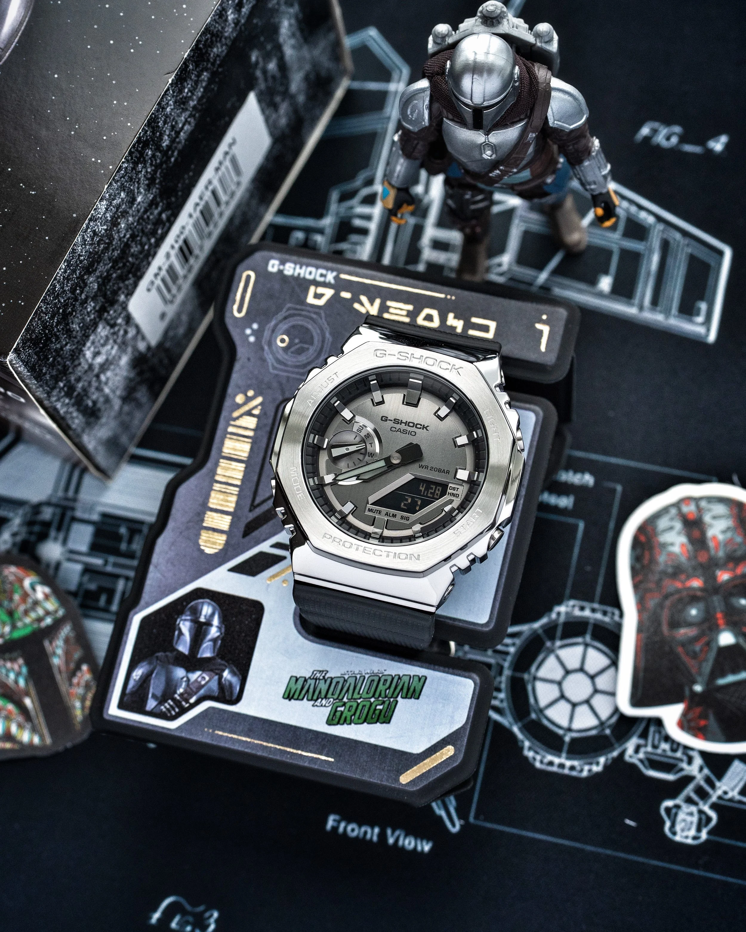 G-SHOCK Mandalorian GM-2100 watch displayed on its Bounty Hunter ID stand with Star Wars themed background and figure