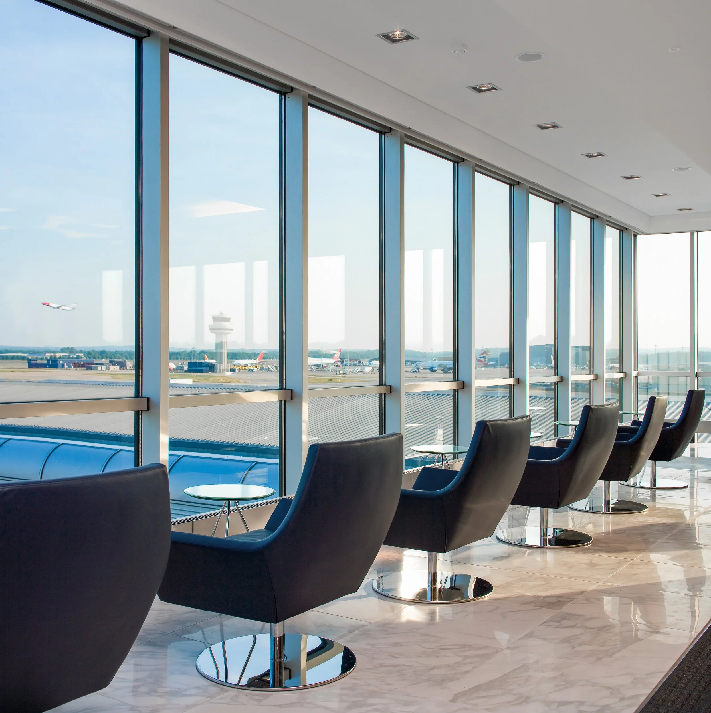 Inside an airport lounge with large windows overlooking the runway, showing chairs and small tables facing the windows.