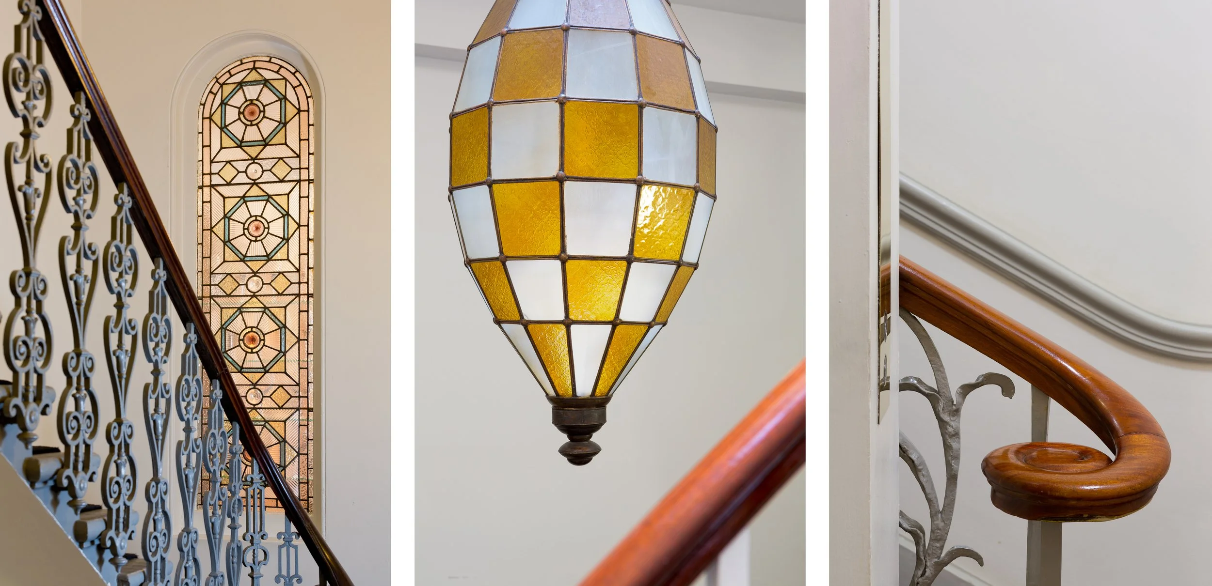 Interior view of a staircase with decorative iron railing, a stained glass window with geometric patterns, and a hanging stained glass pendant light with yellow and white panels.