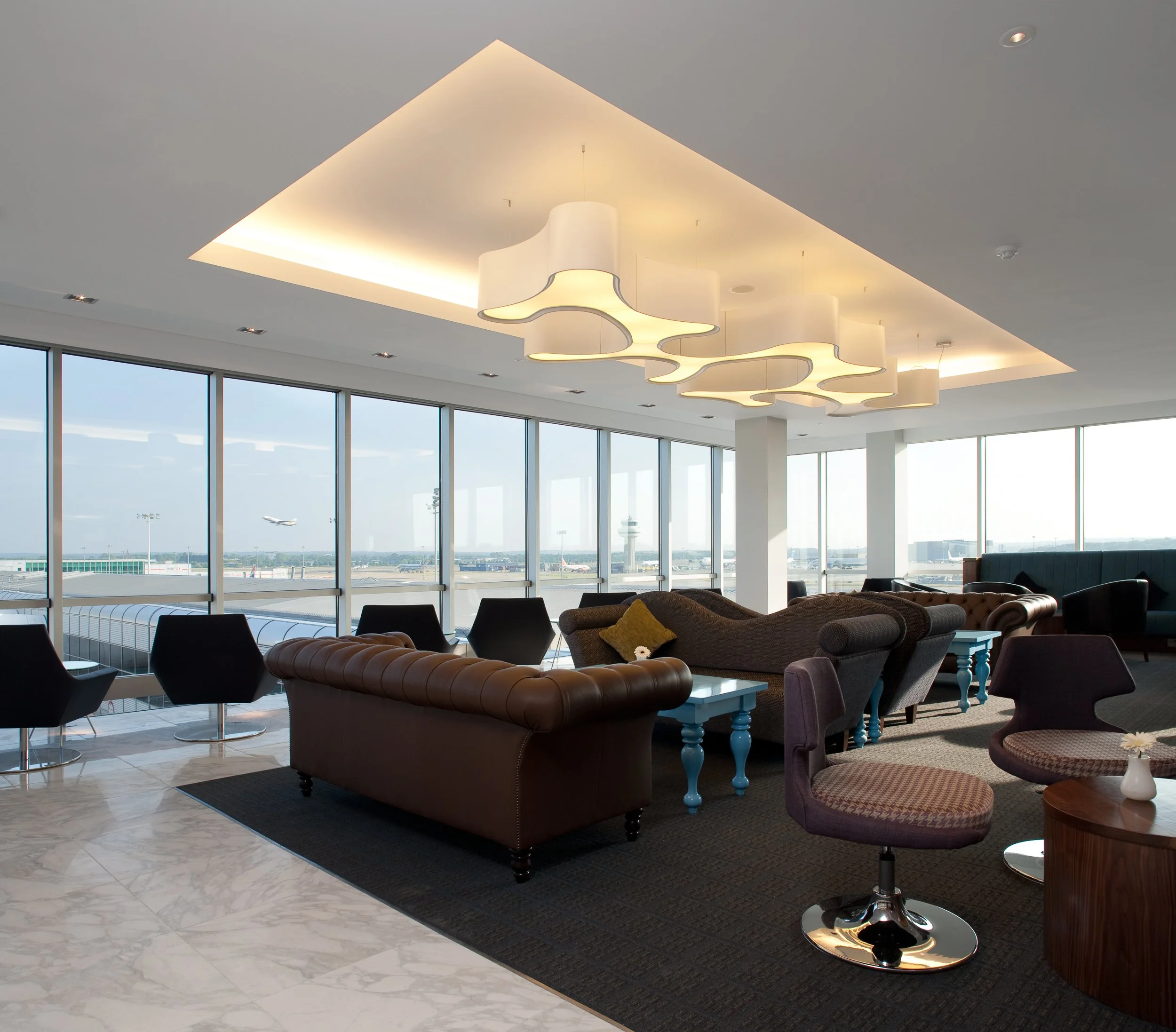 Modern airport lounge with large windows, leather and fabric seating, and a decorative ceiling light fixture.