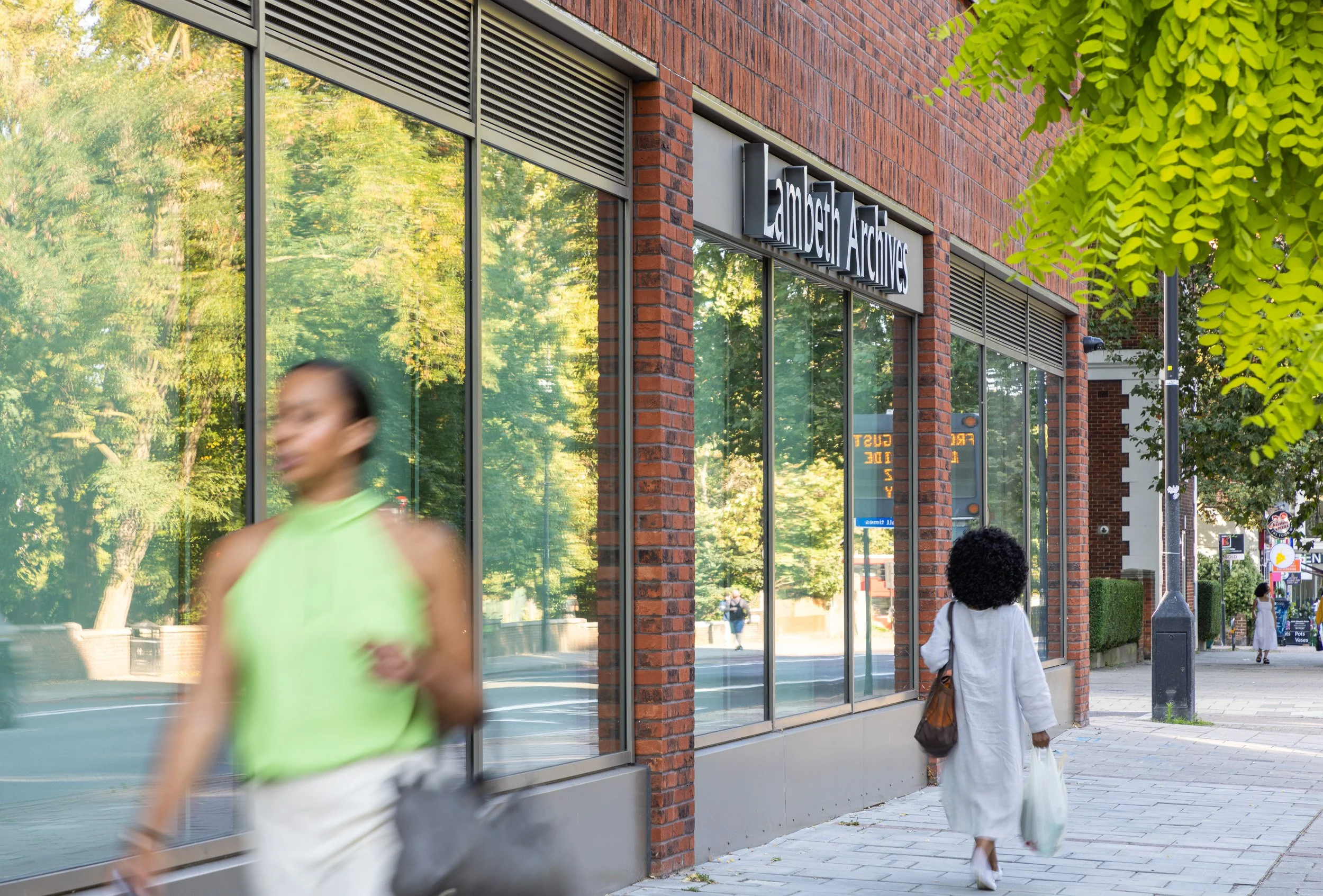 A woman with curly hair wearing a white dress carries shopping bags walking past the Lambeth Archives building with large glass windows reflecting trees, on a sunny day.