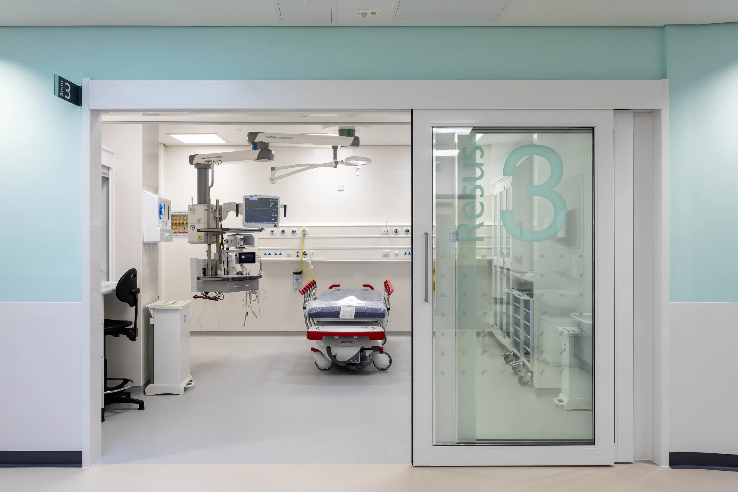 Hospital operating room with an adjustable surgical bed, medical equipment, and monitors, seen through a glass sliding door with '3RD' printed on it.