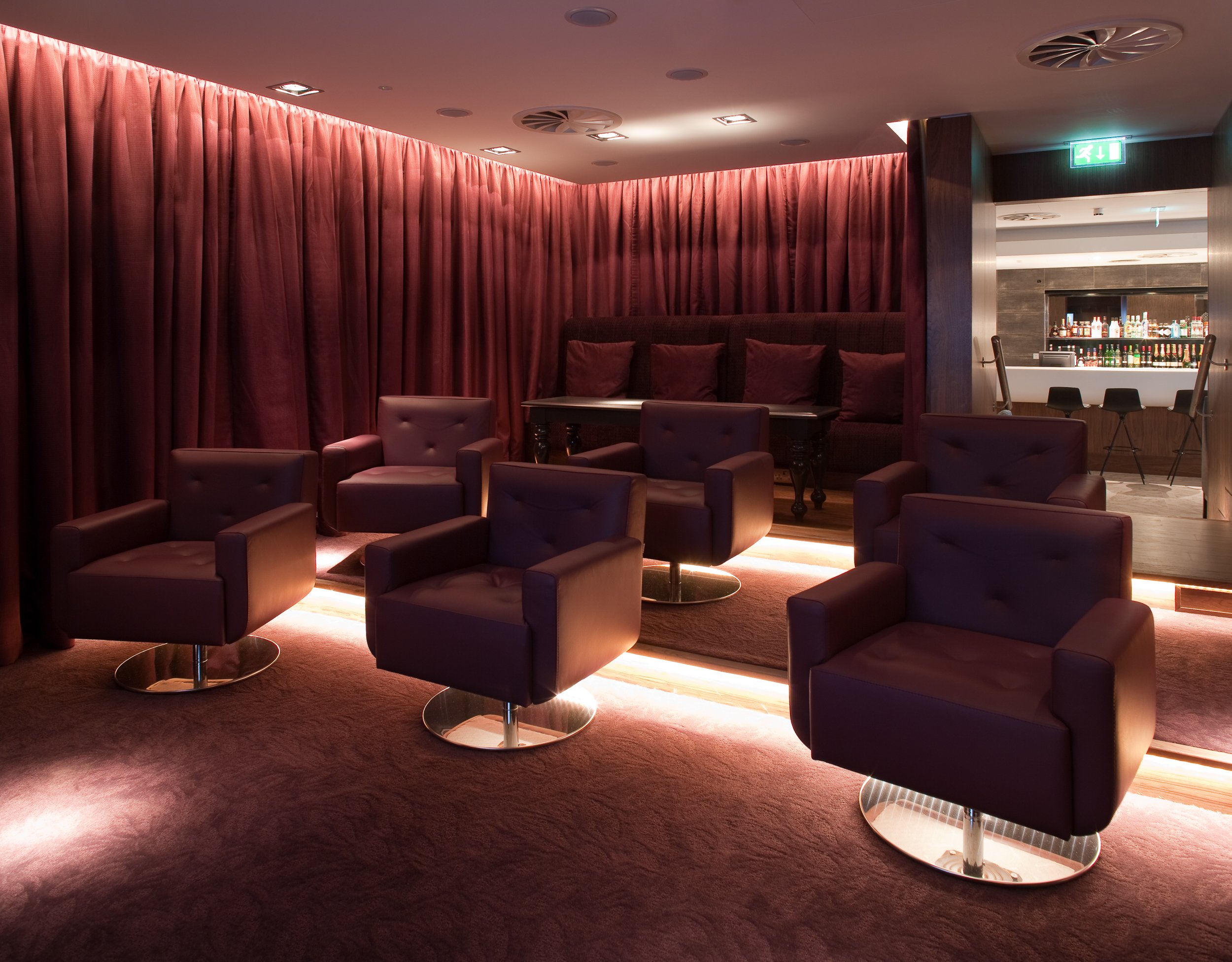 A lounge area with six purple armchairs arranged on a raised platform with underlighting, surrounded by purple curtains and a carpeted floor. In the background, there is a bar with bottles on shelves and a few barstools.