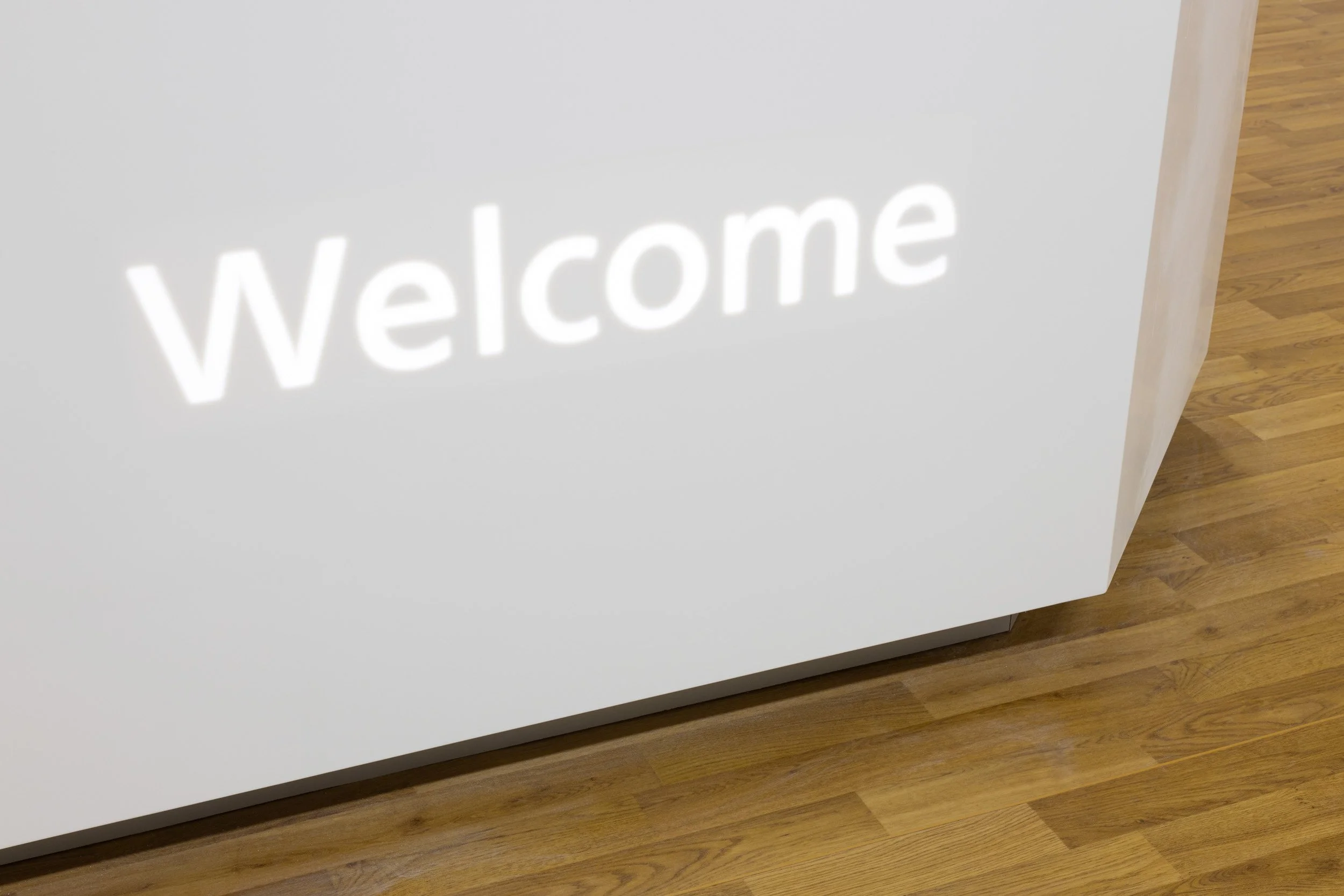A white wall with the word 'Welcome' in illuminated letters, partially visible, next to a wooden floor.