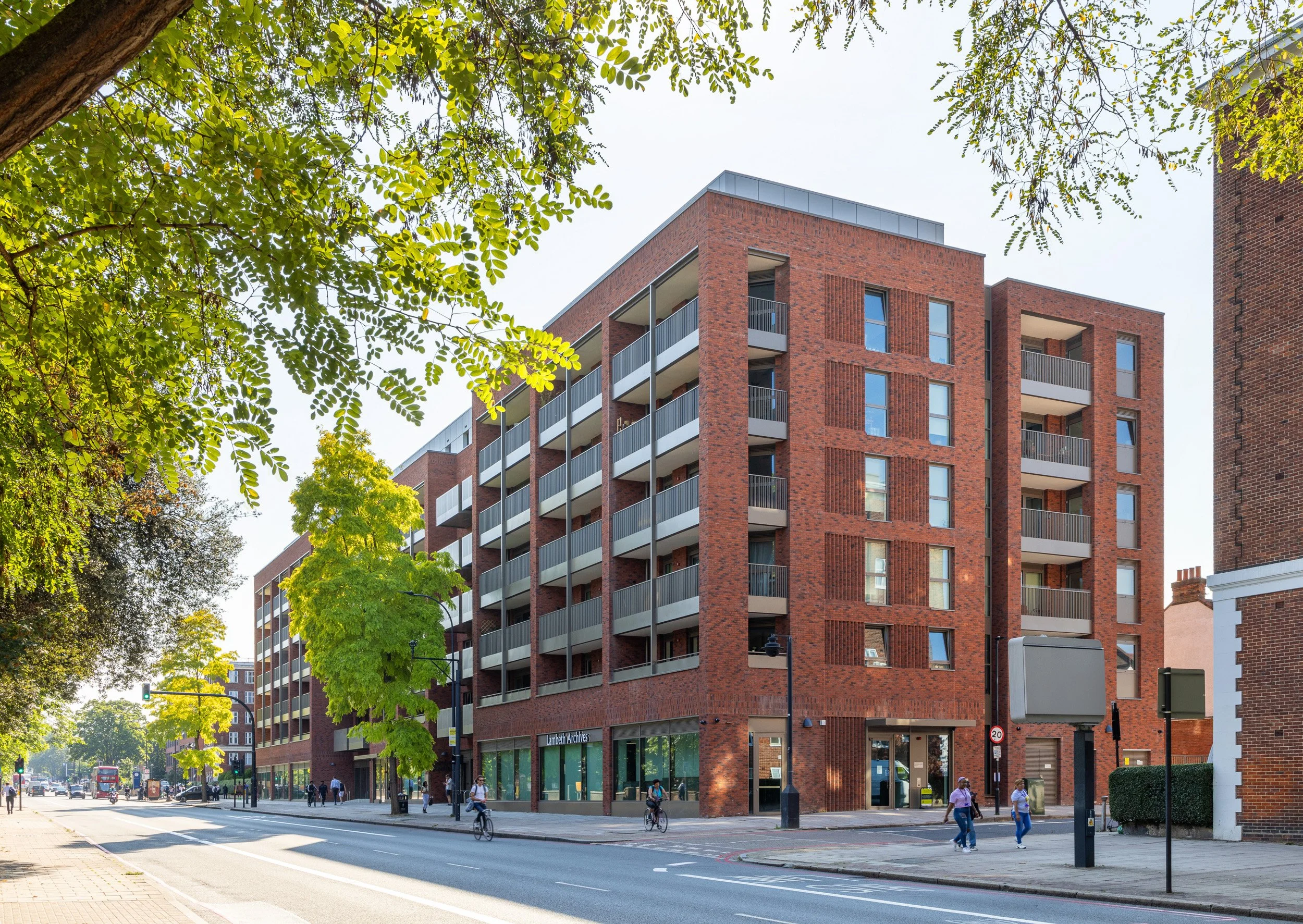 Modern multi-story red brick building on a city street with trees and pedestrians