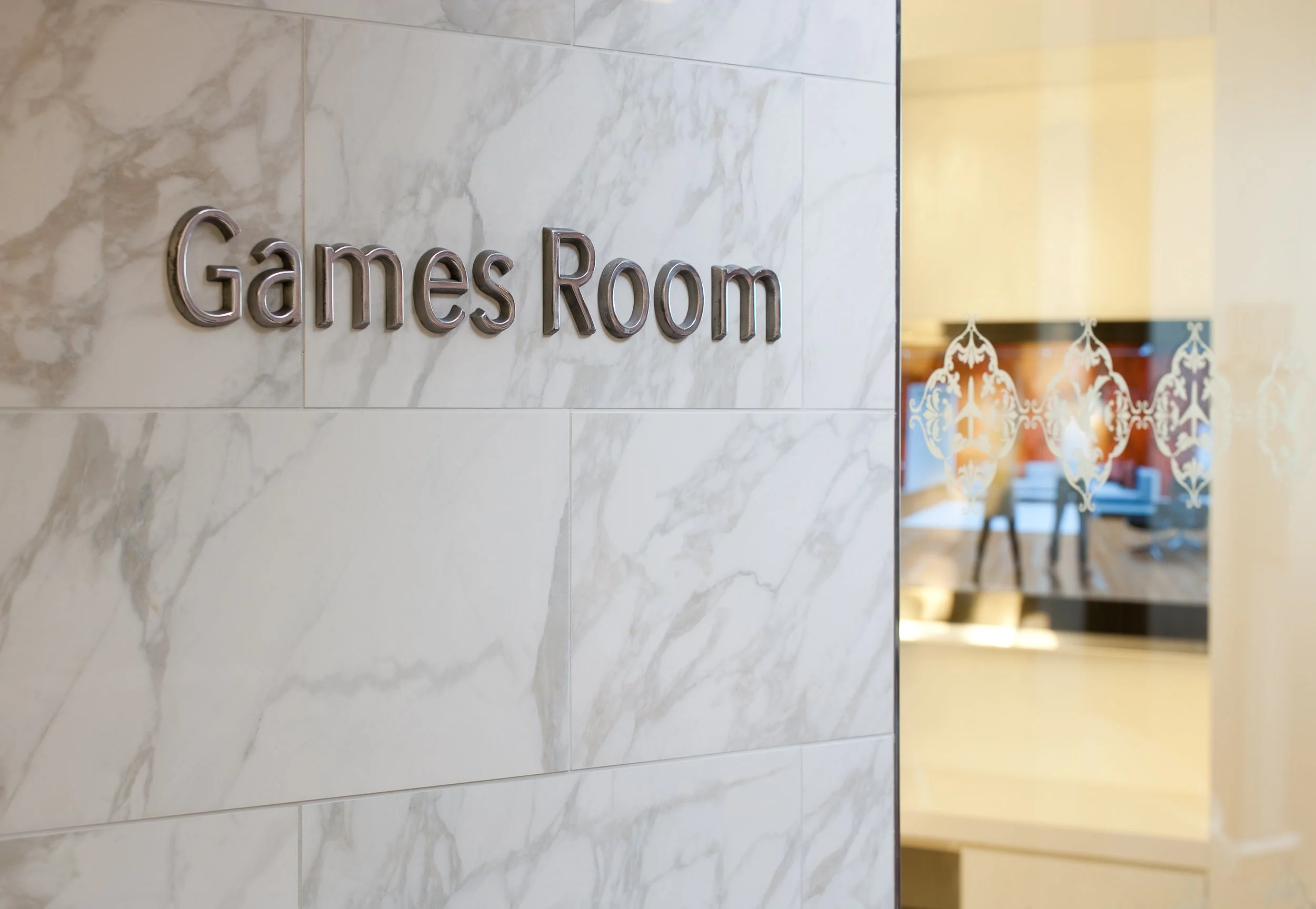 A sign that reads 'Games Room' on a marble wall, with a glass door and a reflection of a living room inside.