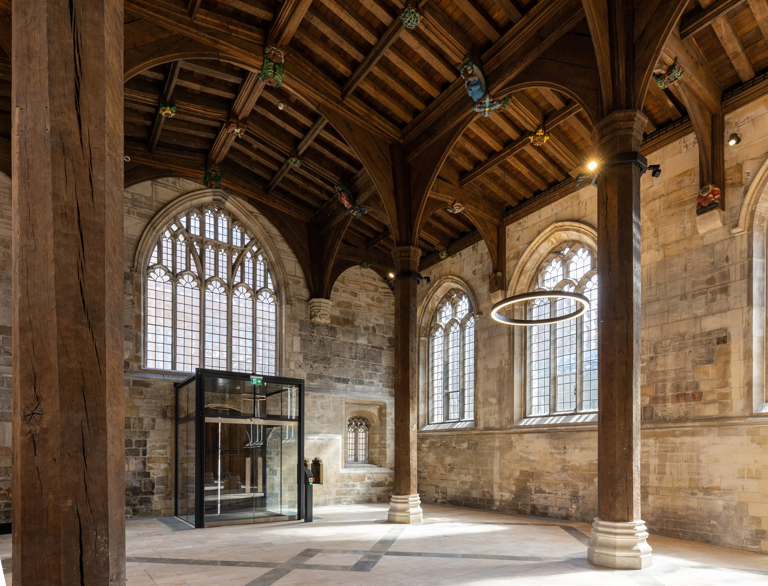 Interior of a historic church with large stained glass windows, wooden ceiling beams, and stone walls. There is a modern glass elevator and a circular light fixture hanging from the ceiling.