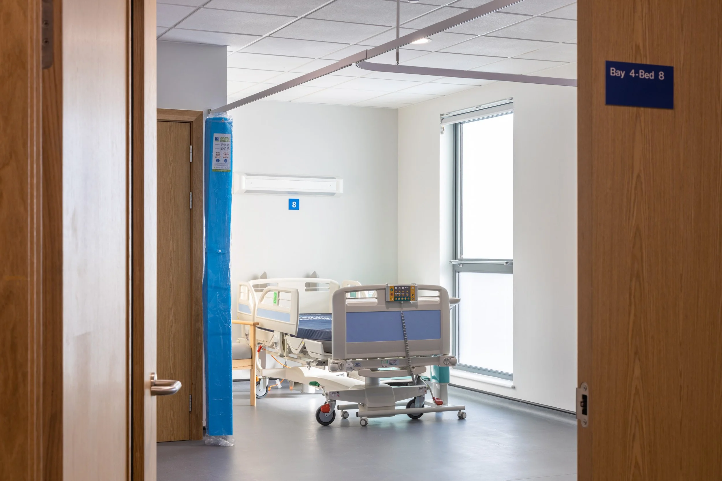 View of a hospital room through an open door, with a hospital bed and a window.