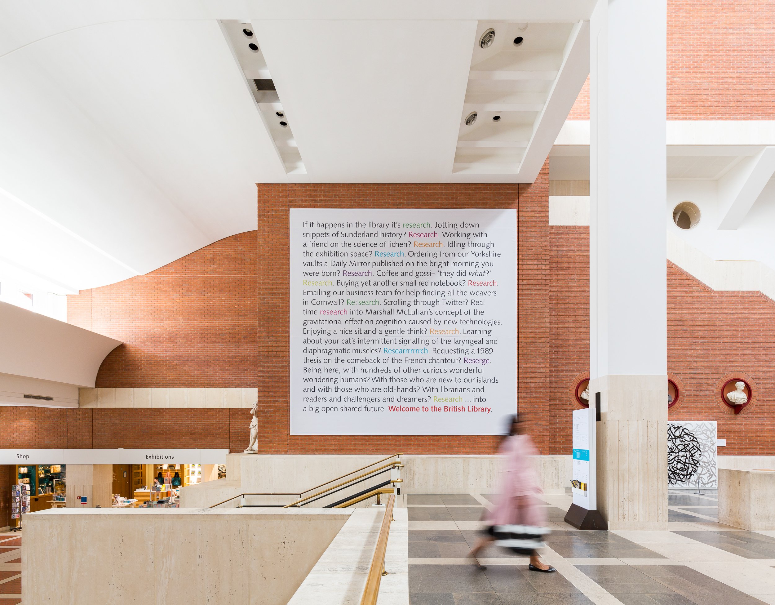 Interior view of a modern library with a brick wall, large informational display, and a blurred person walking past. Signage indicates sections for shop and exhibitions.
