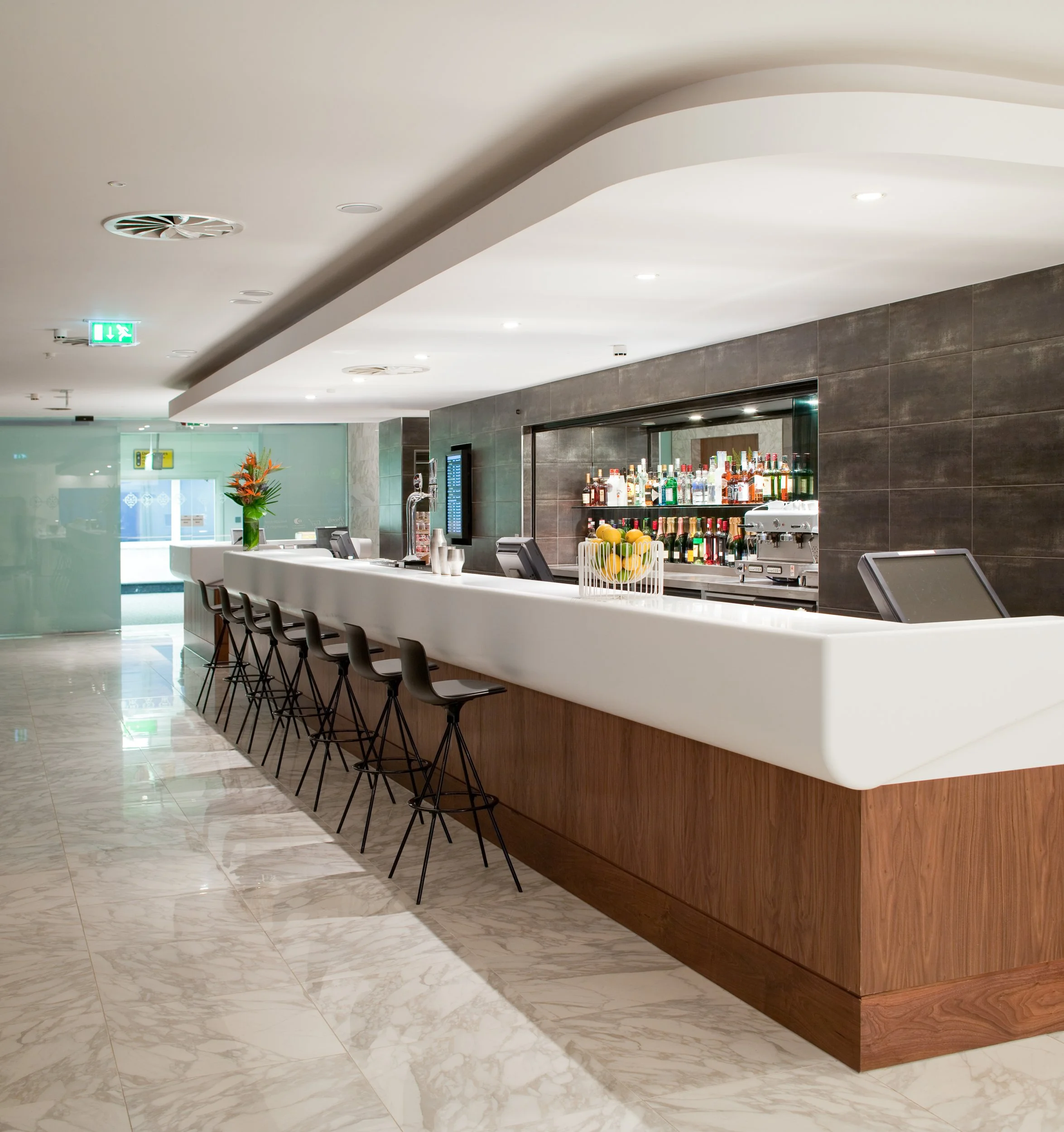Modern hotel bar with a white counter, bar stools, bottles of alcohol on shelves, and a vase with flowers.