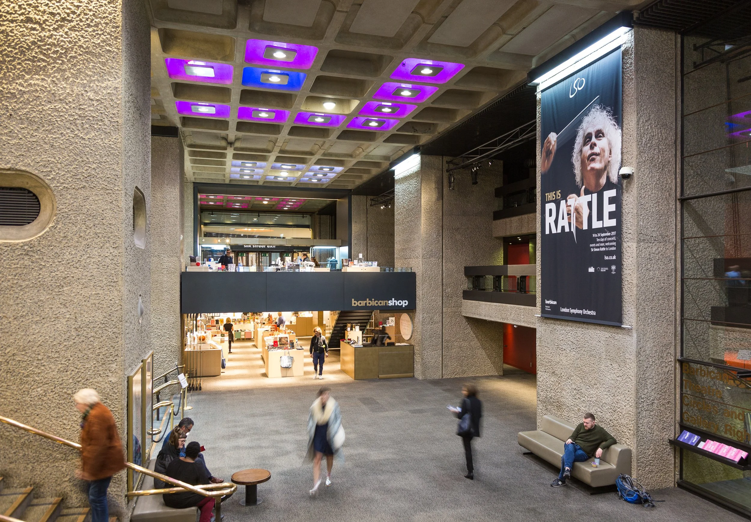 Interior of a public building with people walking and sitting, a large poster of an elderly woman conducting, and a shop named 'barbican shop' on the upper level.
