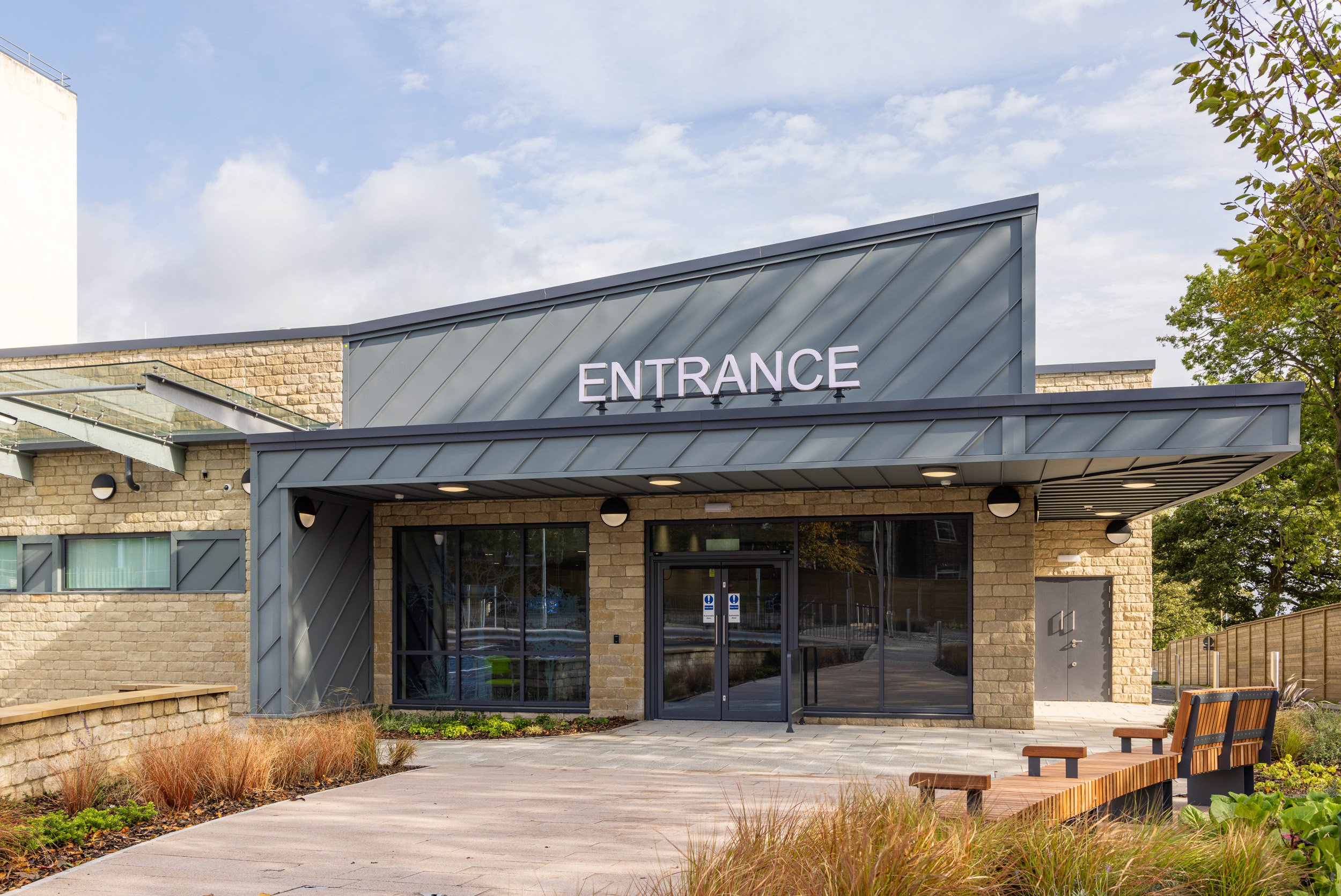 Entrance to a modern building with a metal roof, glass double doors, and a brick facade. There is a bench and landscaped plants outside.