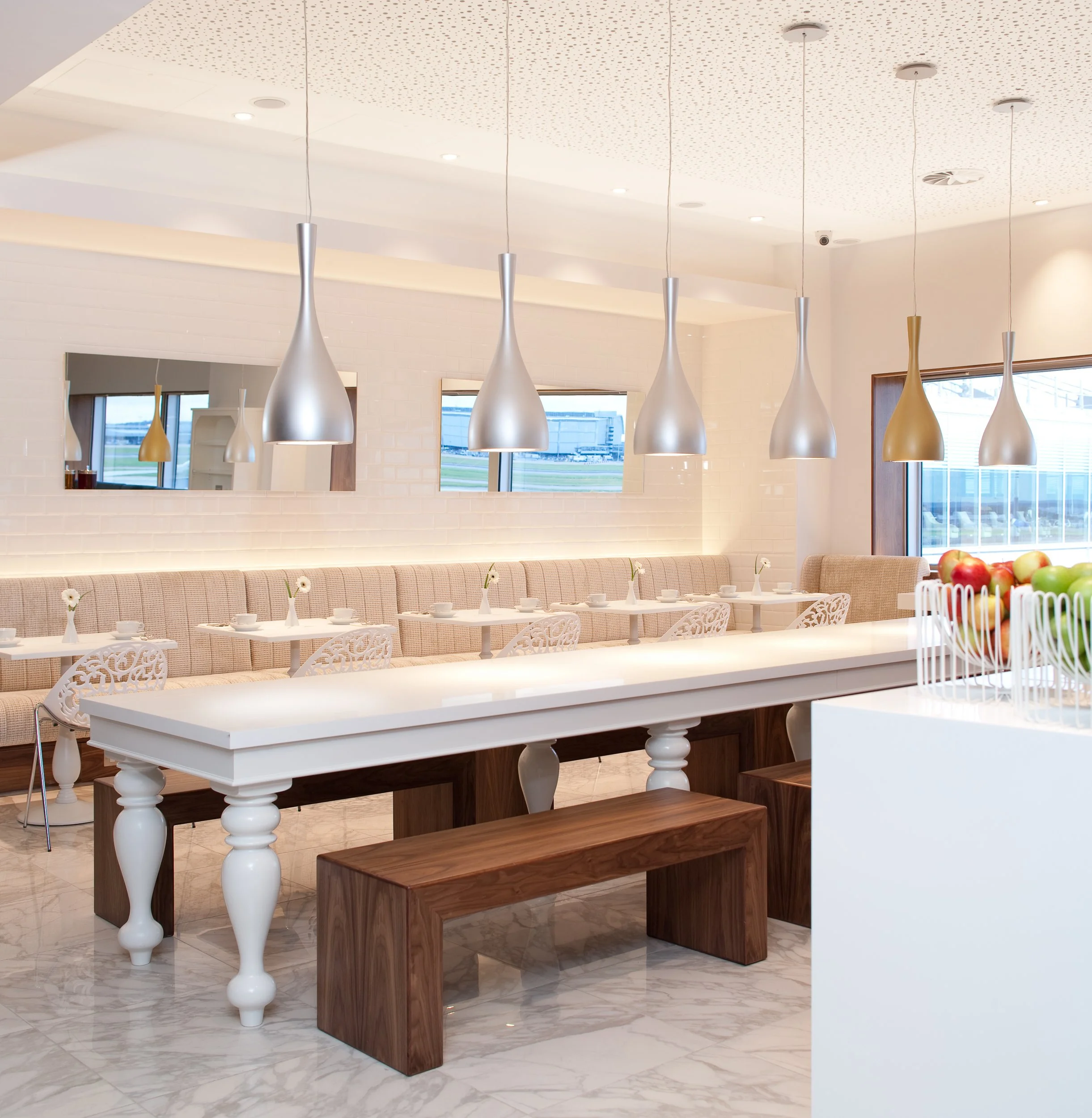 Modern dining area with hanging silver pendant lights, beige upholstered bench seating, white tables set with small flower vases, windows revealing an outside view, and a countertop with bowls of fruit.
