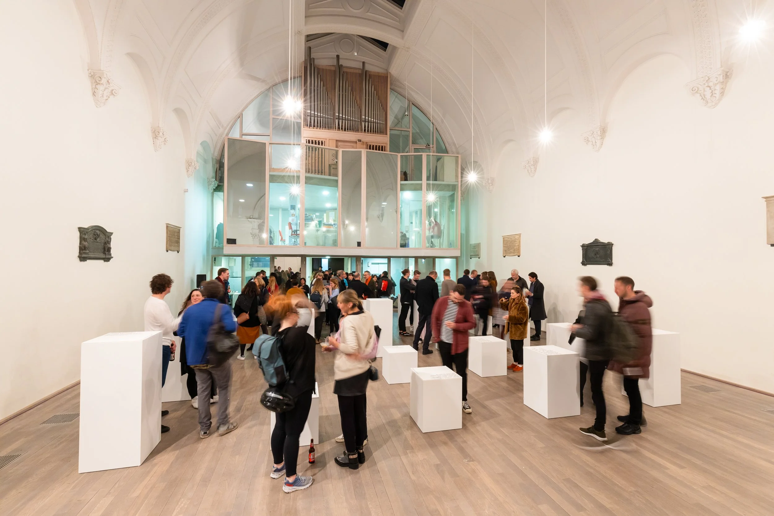 People attending an art exhibition or event inside a large, high-ceilinged gallery with white walls and wooden floors. The crowded room features white pedestals, some with informational plaques, and a glass-enclosed upper level with an organ. Attende