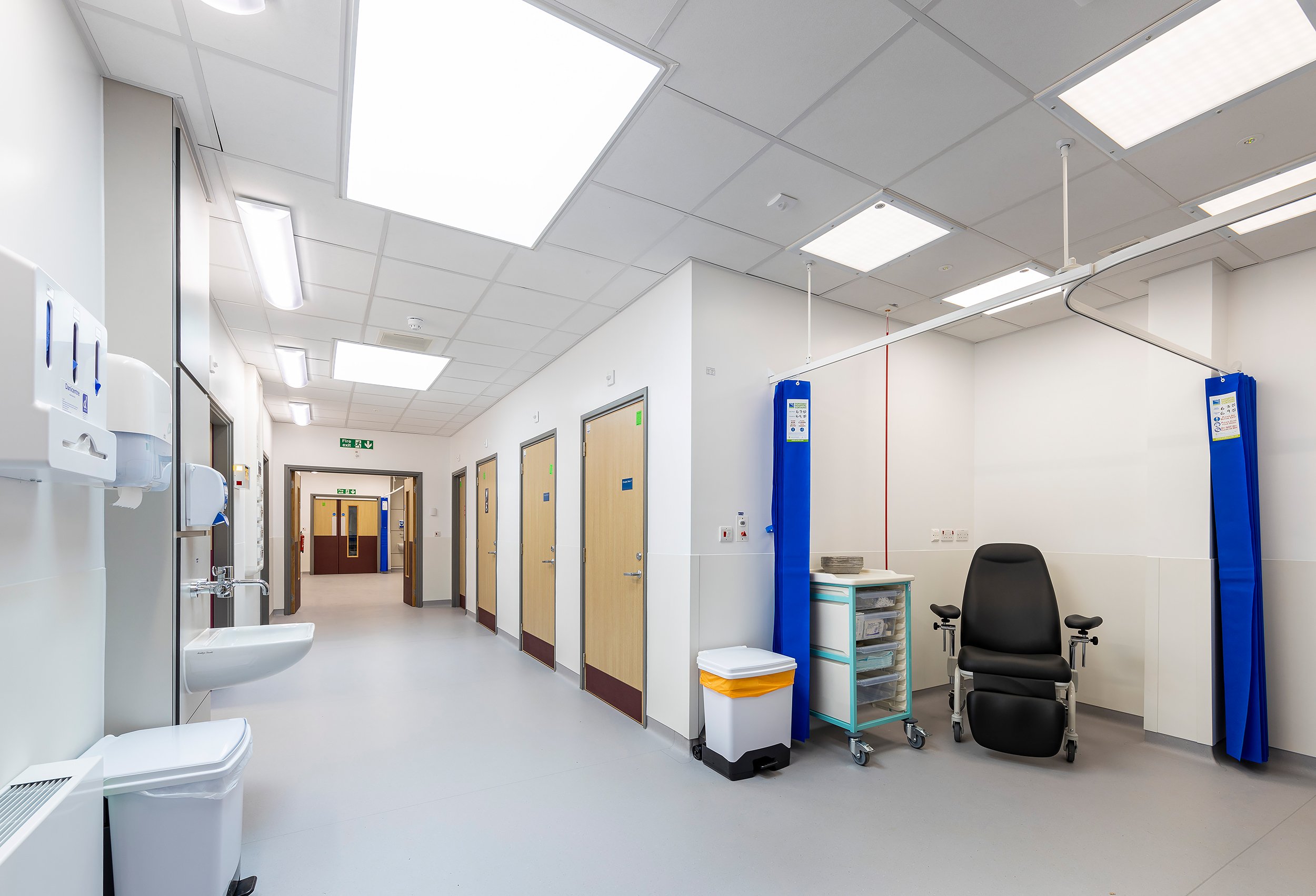 Hospital corridor with multiple patient rooms, a cleaning cart, a chair, medical equipment, and medical gloves dispensers on the wall.