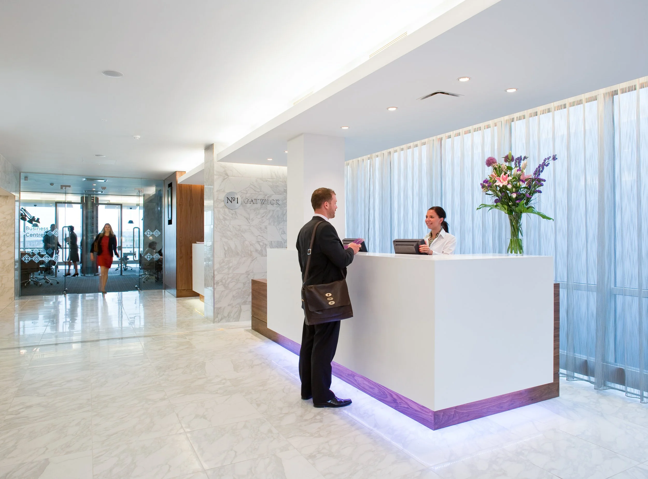 Front desk reception area with a male guest and female receptionist, large flower arrangement, and glass doors leading to the entrance of a hotel or office building.
