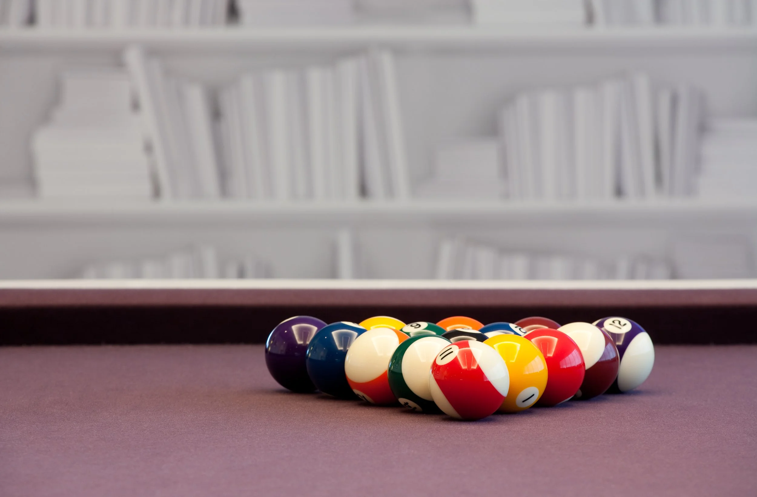Pool balls arranged on a pool table with a blurred white bookshelf in the background.