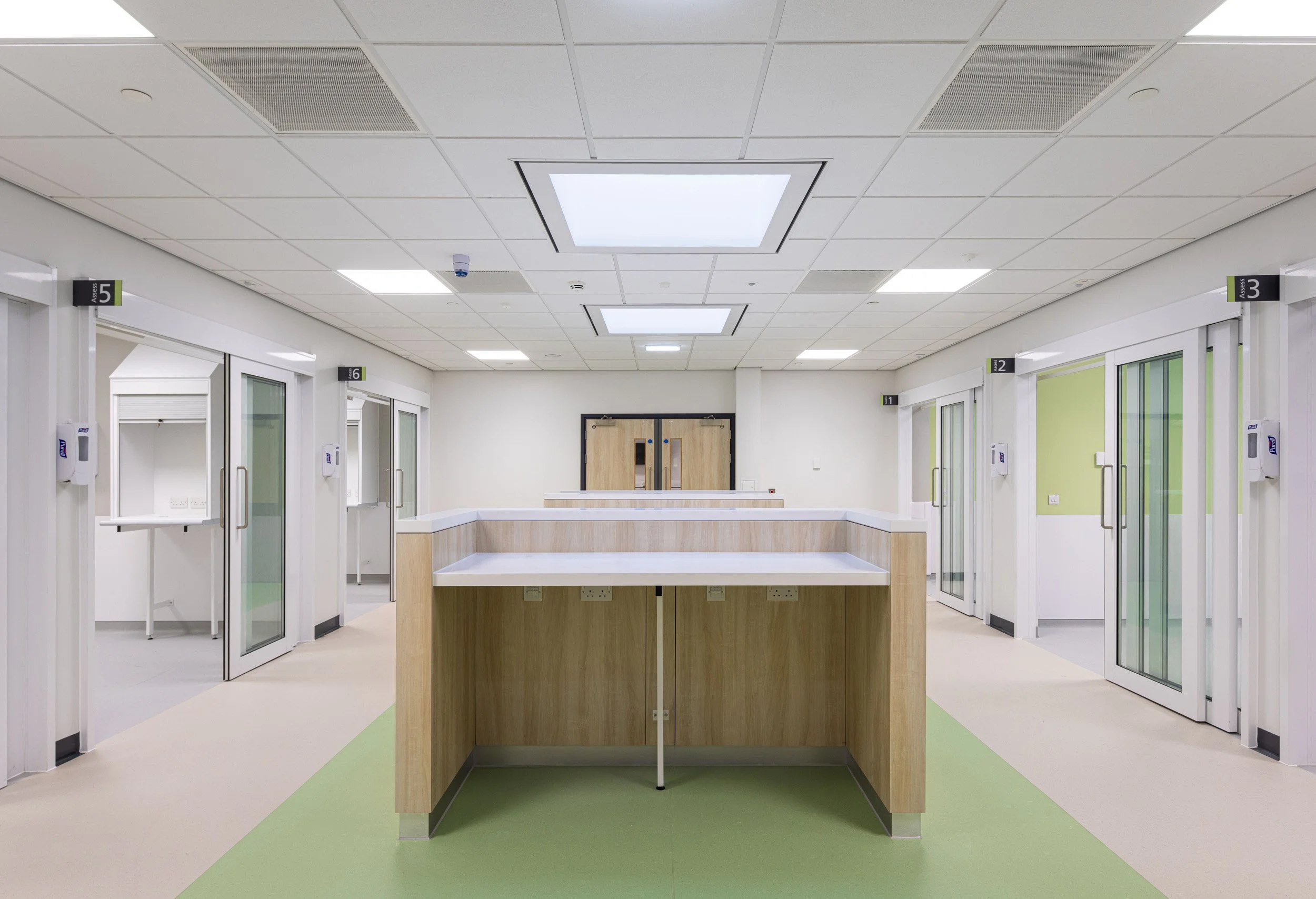 Empty hospital corridor with numbered doors, hand sanitizer dispensers, and a central reception desk, featuring bright ceiling lights and clean, modern design.